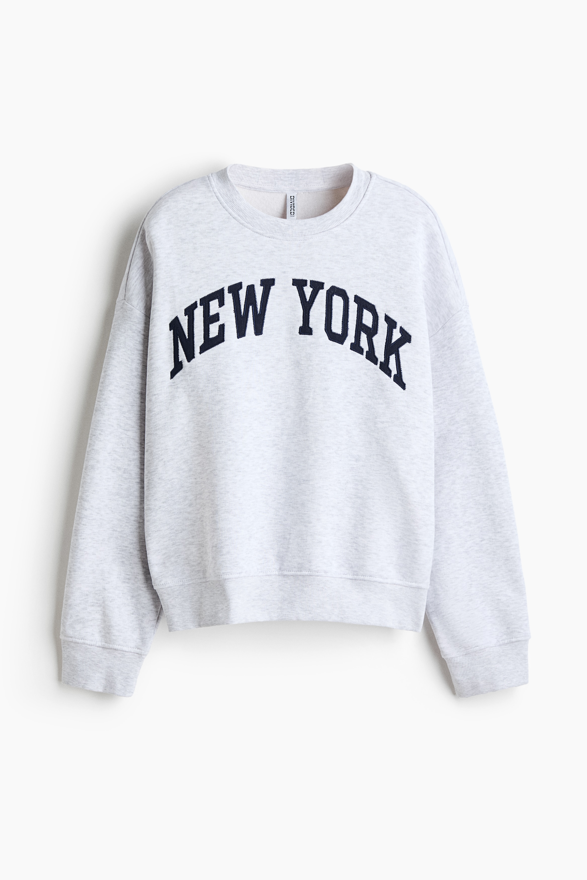 A light grey sweat-fabric sweater featuring a round, rib-trimmed neckline and a dark blue "NEW YORK" printed design across the chest is being worn by the model. The sweater has a relaxed fit with dropped shoulders.