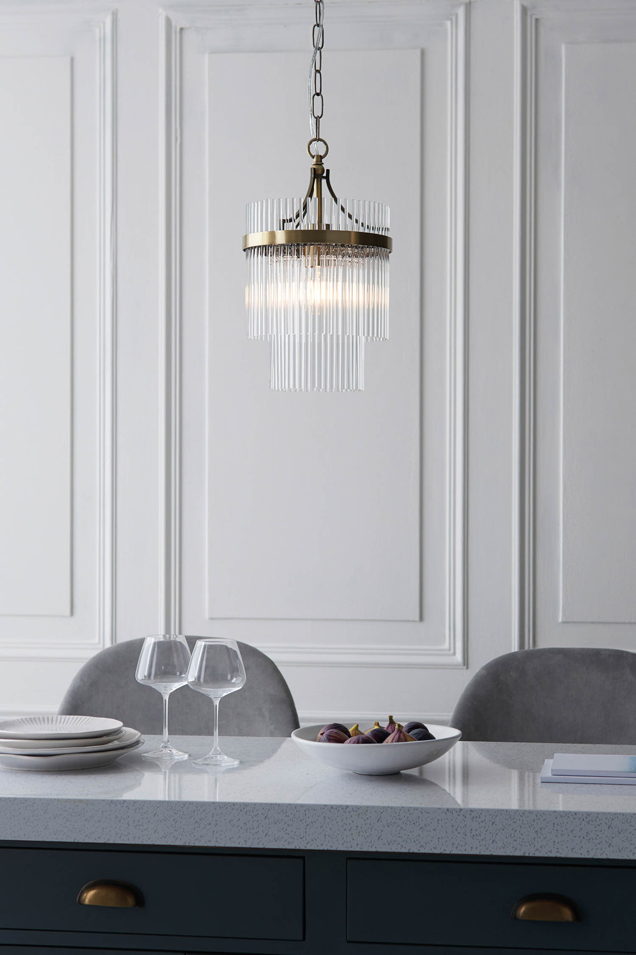 View larger image: A two-tier chandelier is hanging, featuring an antique brass-toned frame supporting two cylindrical layers of vertical clear glass rods. Light is emanating from within, illuminating the rods.