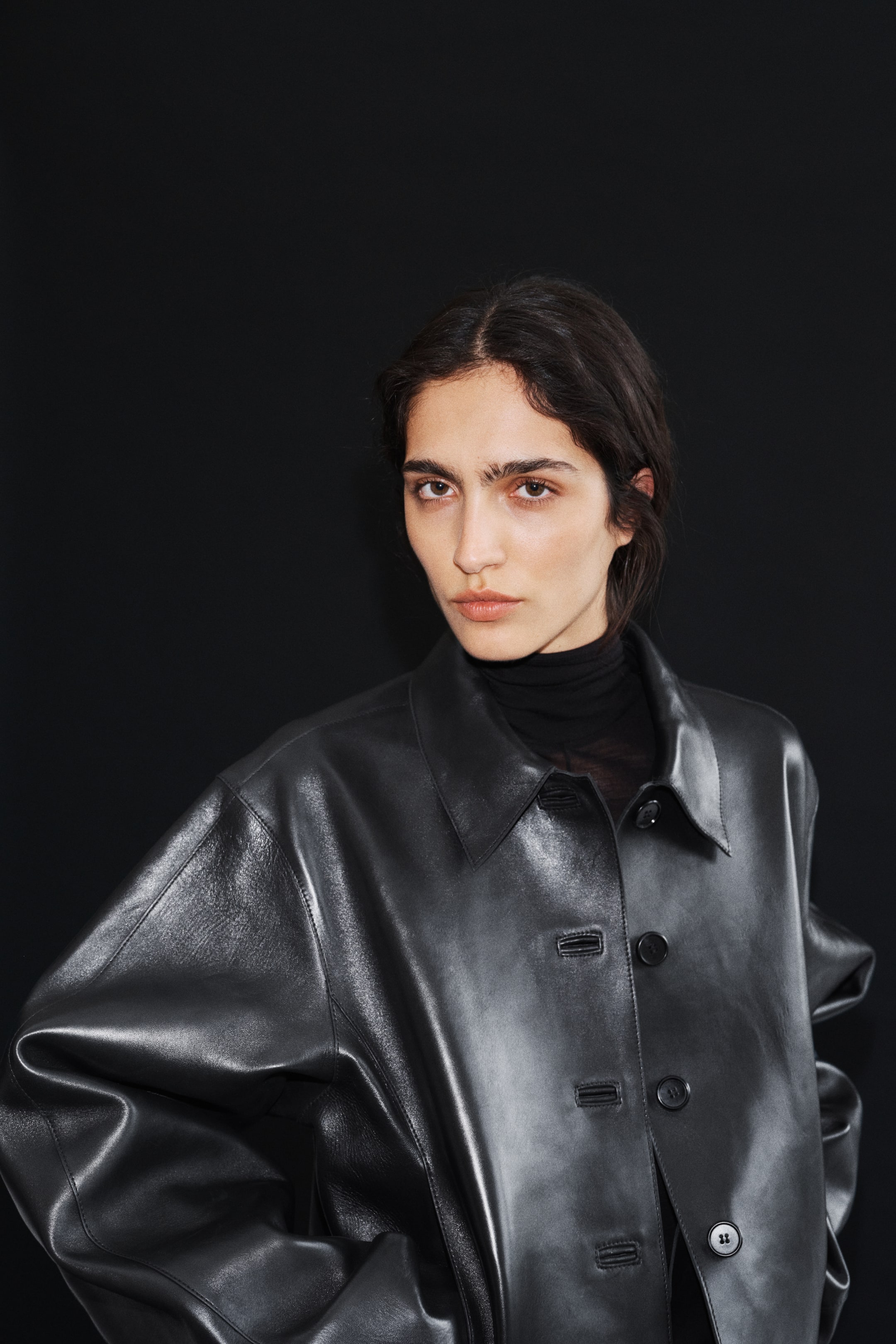 View larger image: The model is wearing a black leather jacket featuring a classic collar and black button-front closure. It is layered over a black turtleneck, with the relaxed fit creating visible folds in the material.