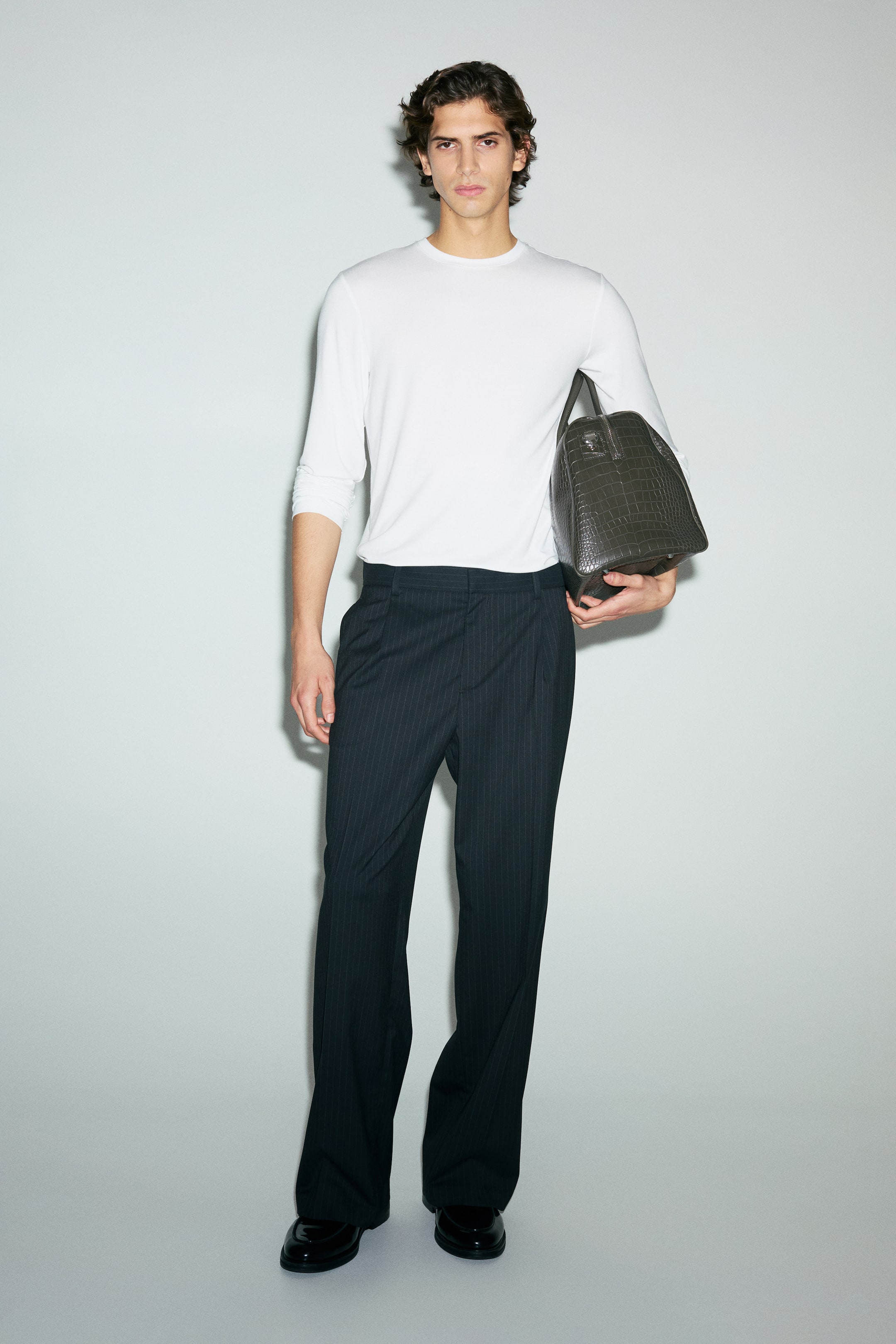 Se større billede: En hvid langærmet t-shirt med rund hals, mørke nålestribede plisserede bukser med brede ben, og sorte polerede loafers præsenteres. En mørk håndtaske med alligatorprægning bæres også, hvilket viser dens strukturerede for- og sideprofil.