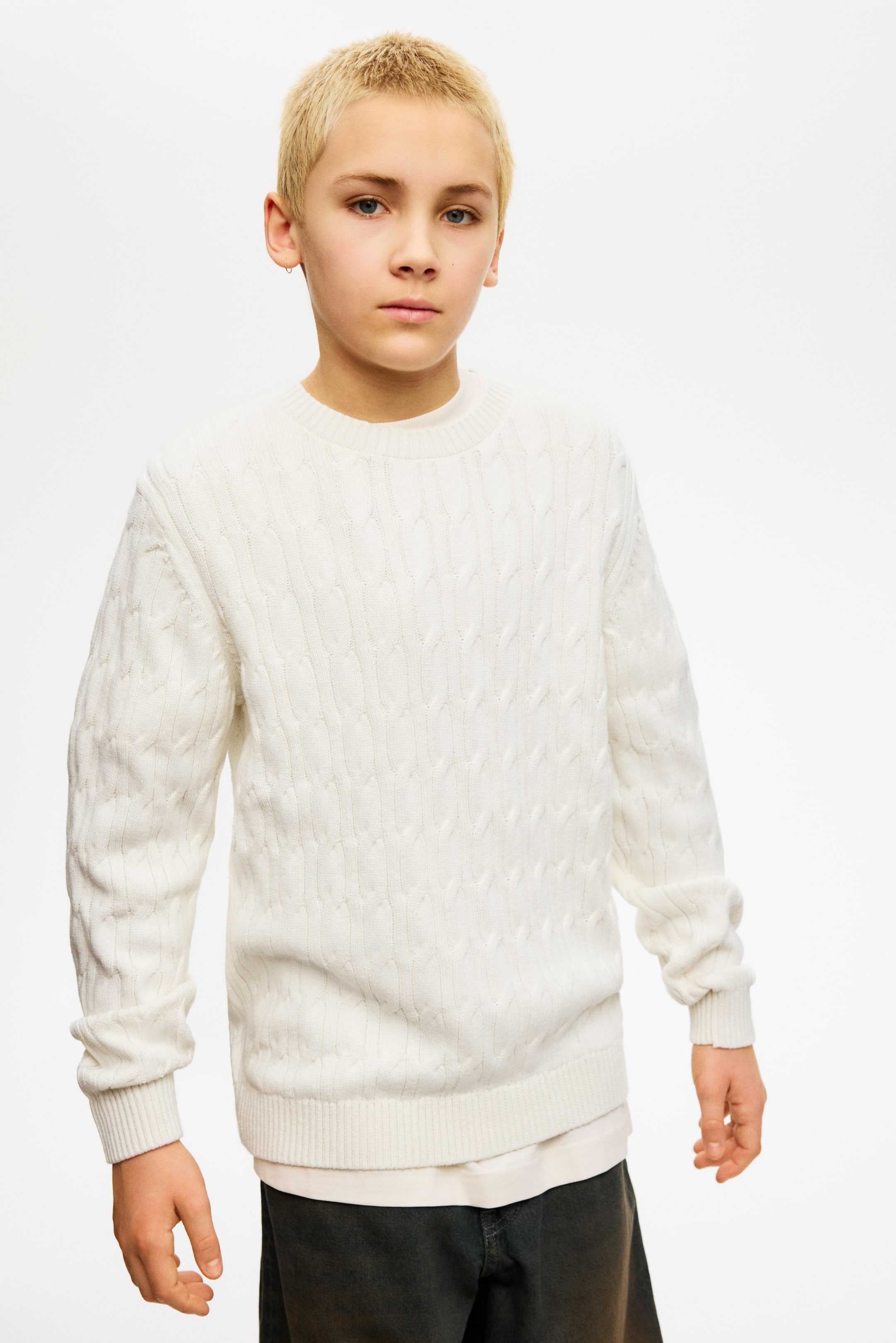 Agrandir l'image: Un pull en maille torsadée blanc est présenté de face, avec un col rond, des manches longues avec des poignets côtelés et un ourlet côtelé. Un t-shirt blanc est visible en dessous, dépassant sous l'ourlet du pull.