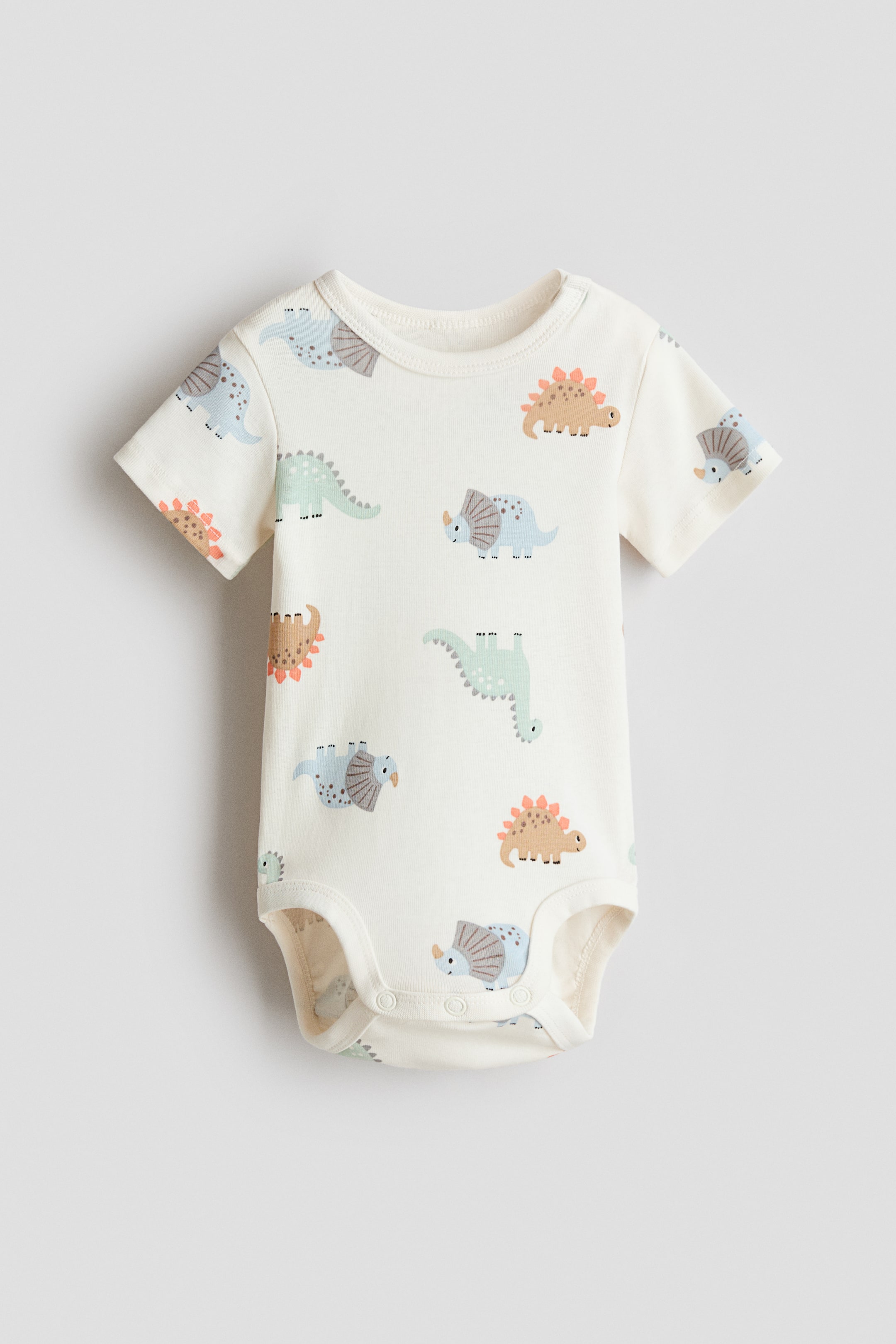 Xem ảnh lớn hơn: Một bộ áo liền quần cho trẻ sơ sinh màu kem hướng MẶT TRƯỚC, với tay ngắn, cổ áo tròn có đường viền và các họa tiết khủng long hoạt hình trải khắp vải. Khóa bấm có thể thấy ở phần đũng quần.
