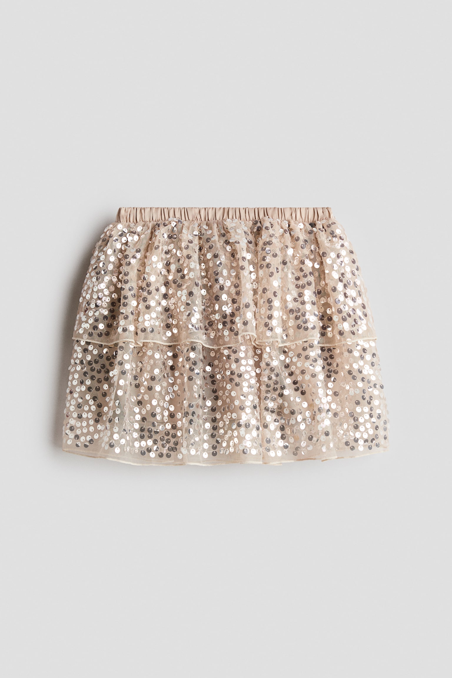 A light beige, tiered skirt is shown from the front, featuring an elasticized satin waistband and two ruffled layers of sheer tulle embellished with all-over silver sequins.