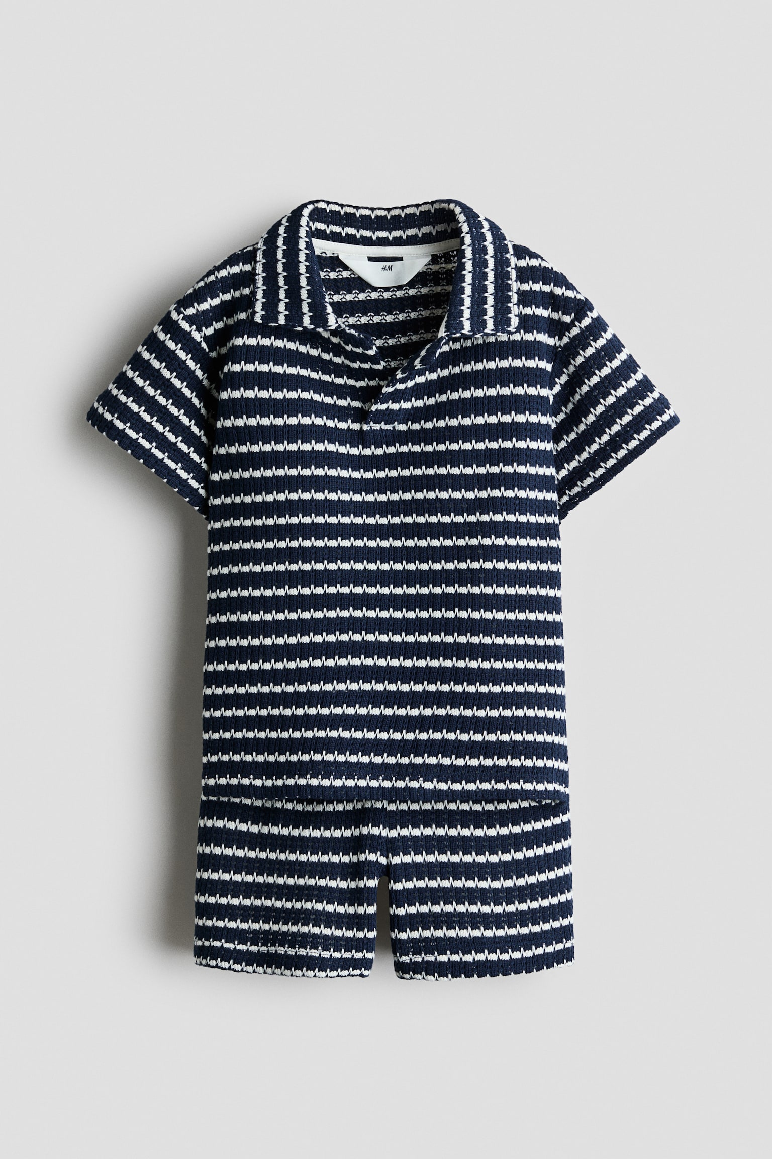 A matching two-piece knit set is shown from the front, featuring a navy and white horizontal zig-zag stripe pattern. The set includes a short-sleeved polo shirt with an open collar and matching shorts.