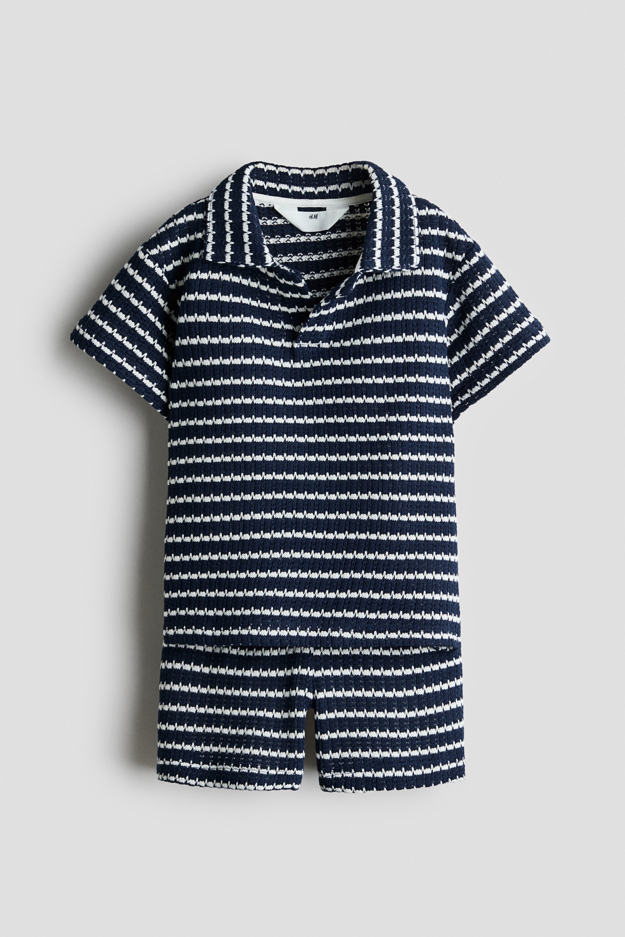 Ampliar la imagen: Se muestra de frente un conjunto tejido de dos piezas a juego, con un patrón de franjas horizontales en zigzag en azul marino y blanco. El conjunto incluye una playera tipo polo de manga corta con cuello abierto y shorts a juego.