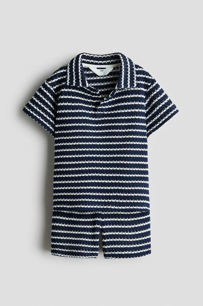 Se muestra de frente un conjunto tejido de dos piezas a juego, con un patrÃ³n de franjas horizontales en zigzag en azul marino y blanco. El conjunto incluye una playera tipo polo de manga corta con cuello abierto y shorts a juego. - 1