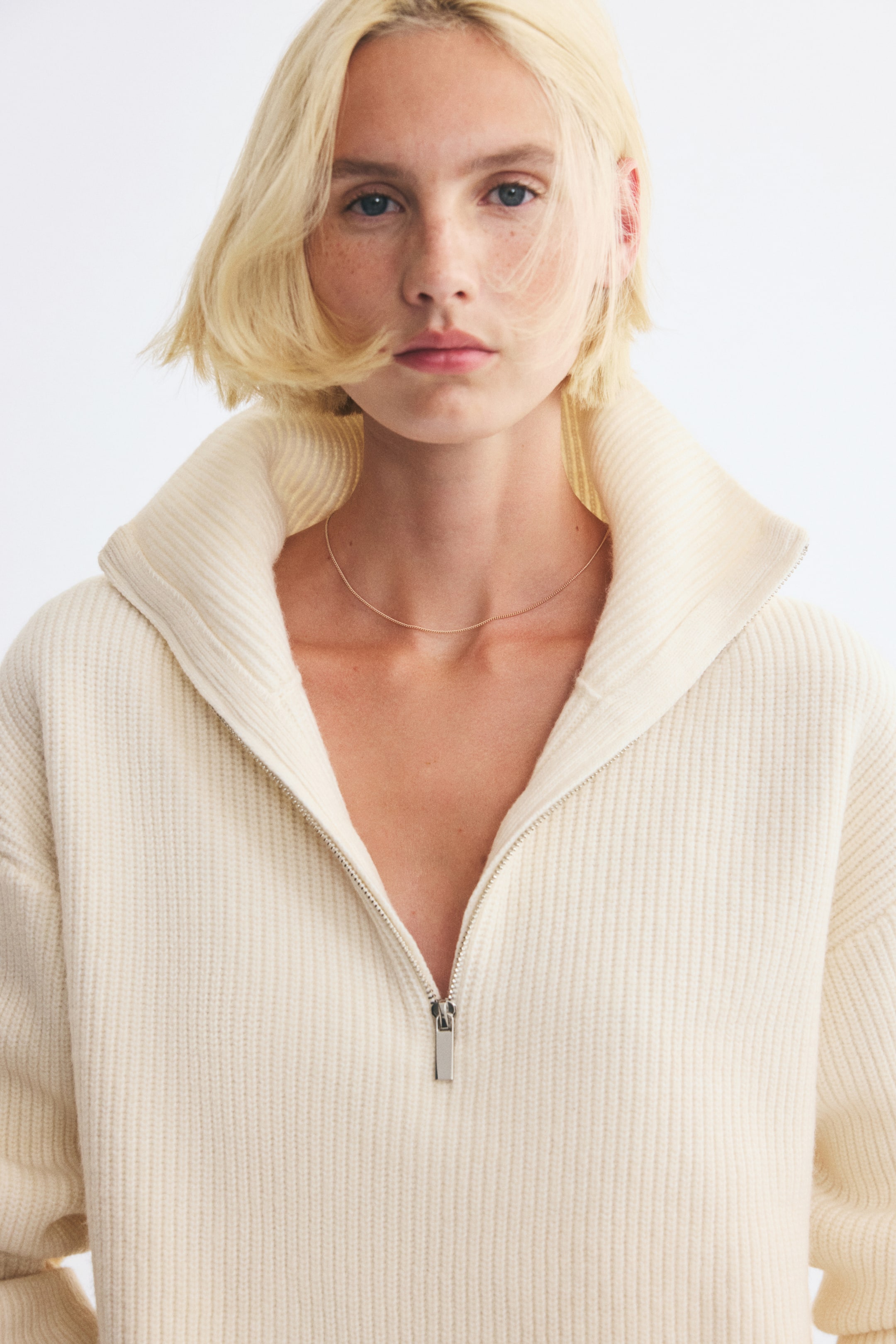 Größeres Bild anzeigen: Ein cremefarbener Pullover im Englischen Rippenstrick mit hohem Kragen und halbem Reißverschluss wird von dem Model getragen. Der silberfarbene Reißverschluss ist teilweise geöffnet und setzt den breiten Kragen sowie die überschnittenen Schultern in Szene.