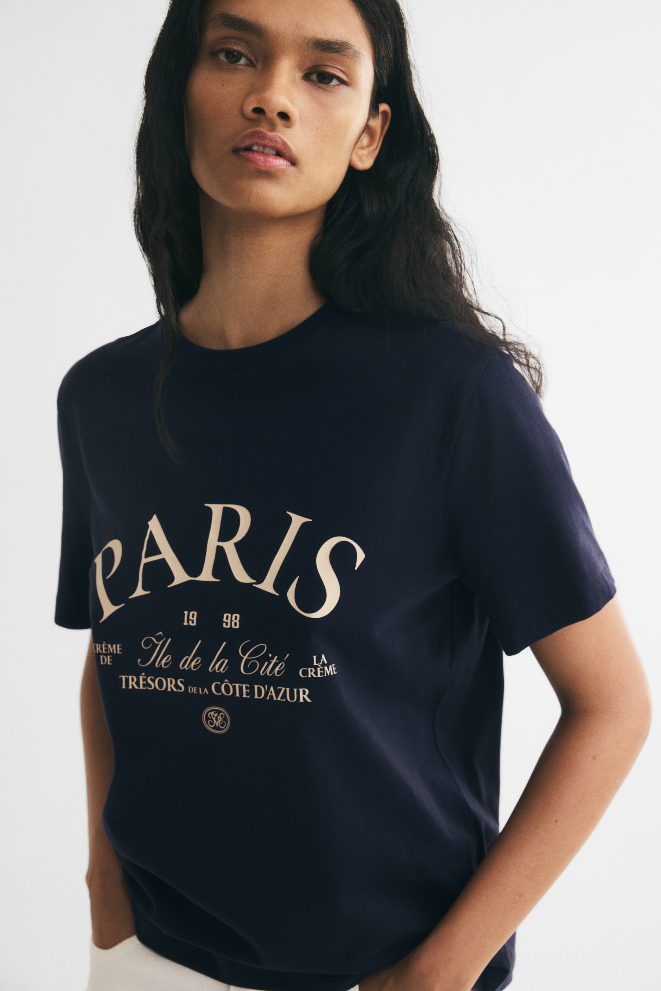 View larger image: A dark blue jersey T-shirt with a round neckline and a cream-coloured graphic print featuring the word "PARIS" on the front is worn by the model. The tee has short sleeves with gently dropped shoulders and is styled with light-coloured trousers.