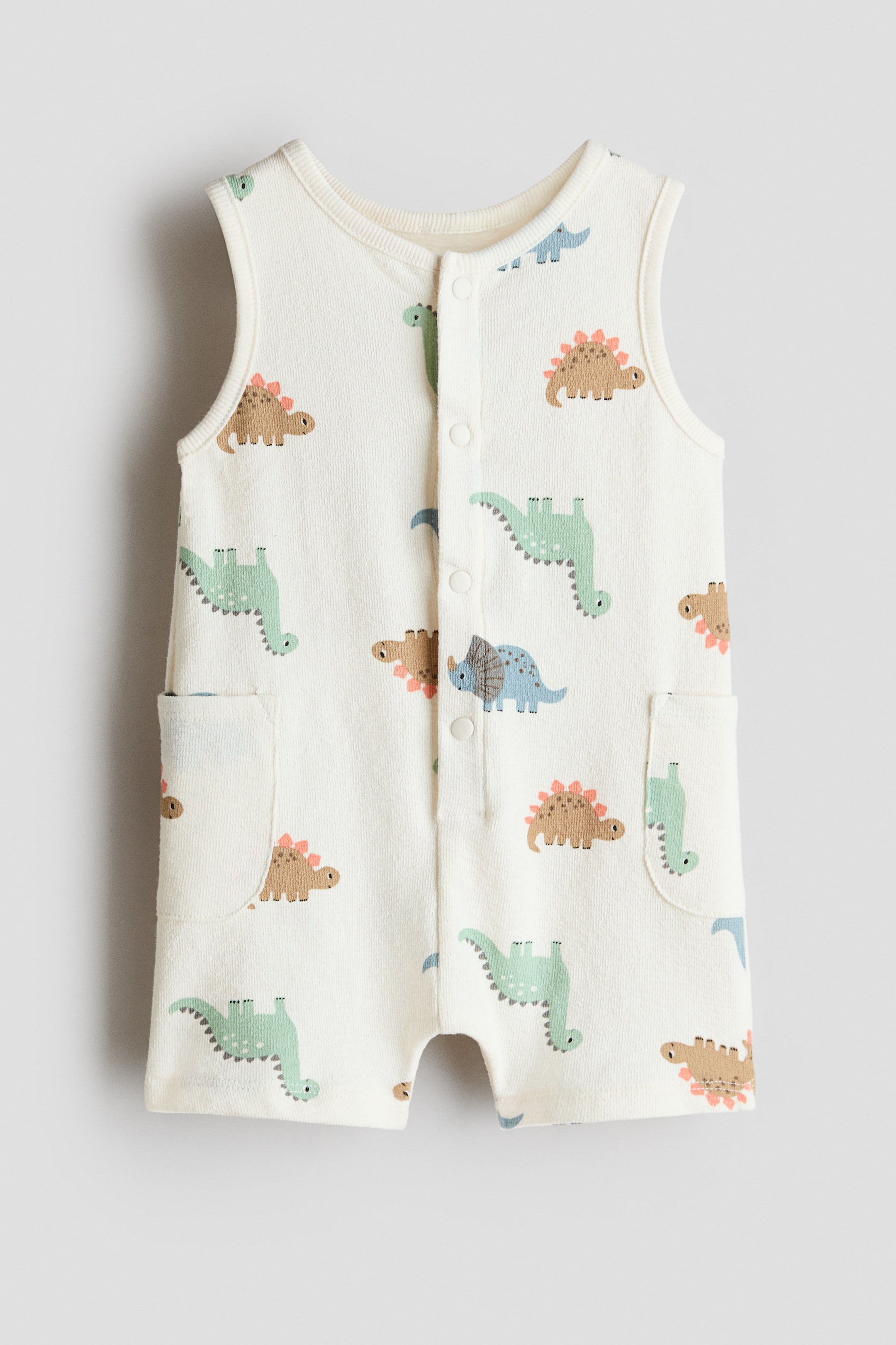 View larger image: Sleeveless romper suit, front view. Crafted from cream cotton terry fabric, it boasts an array of vibrant dinosaur patterns. The design features a round neckline, press-studs along the front, two side patch pockets, and short legs.