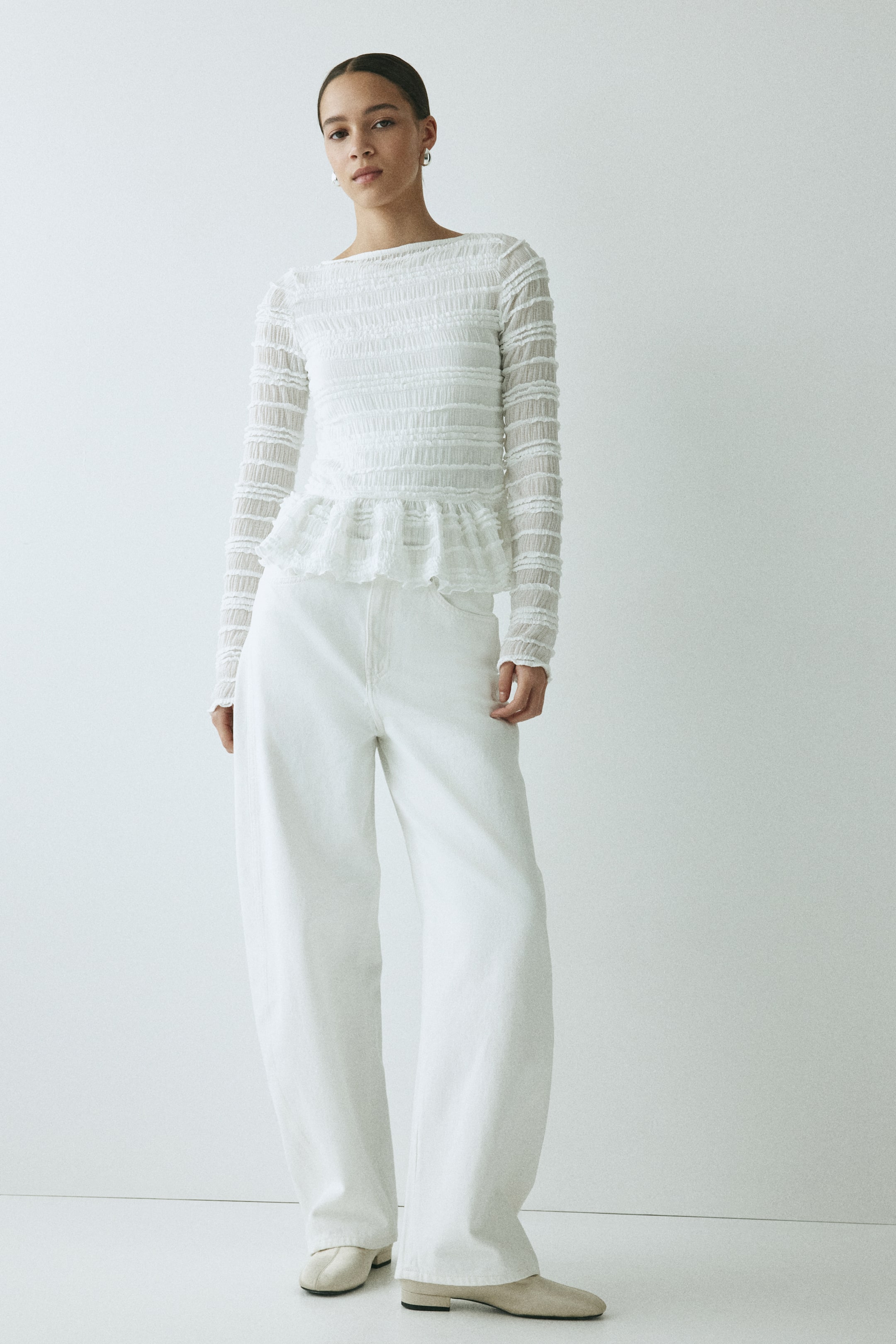Zobraziť väčší obrázok: Biely top s dlhými rukávmi má člnkový výstrih a horizontálne nariasenie, ktoré vytvára textúrovaný efekt na živôtiku i rukávoch, a následne sa rozširuje do volánového peplum lemu. Je kombinovaný s bielymi širokými nohavicami, čím dopĺňa monochromatický outfit.