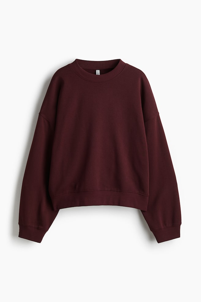 Una felpa color bordeaux è presentata dalla parte anteriore, con scollo rotondo e spalle cadenti. Ha maniche lunghe con polsini a coste e un orlo a coste. - 1