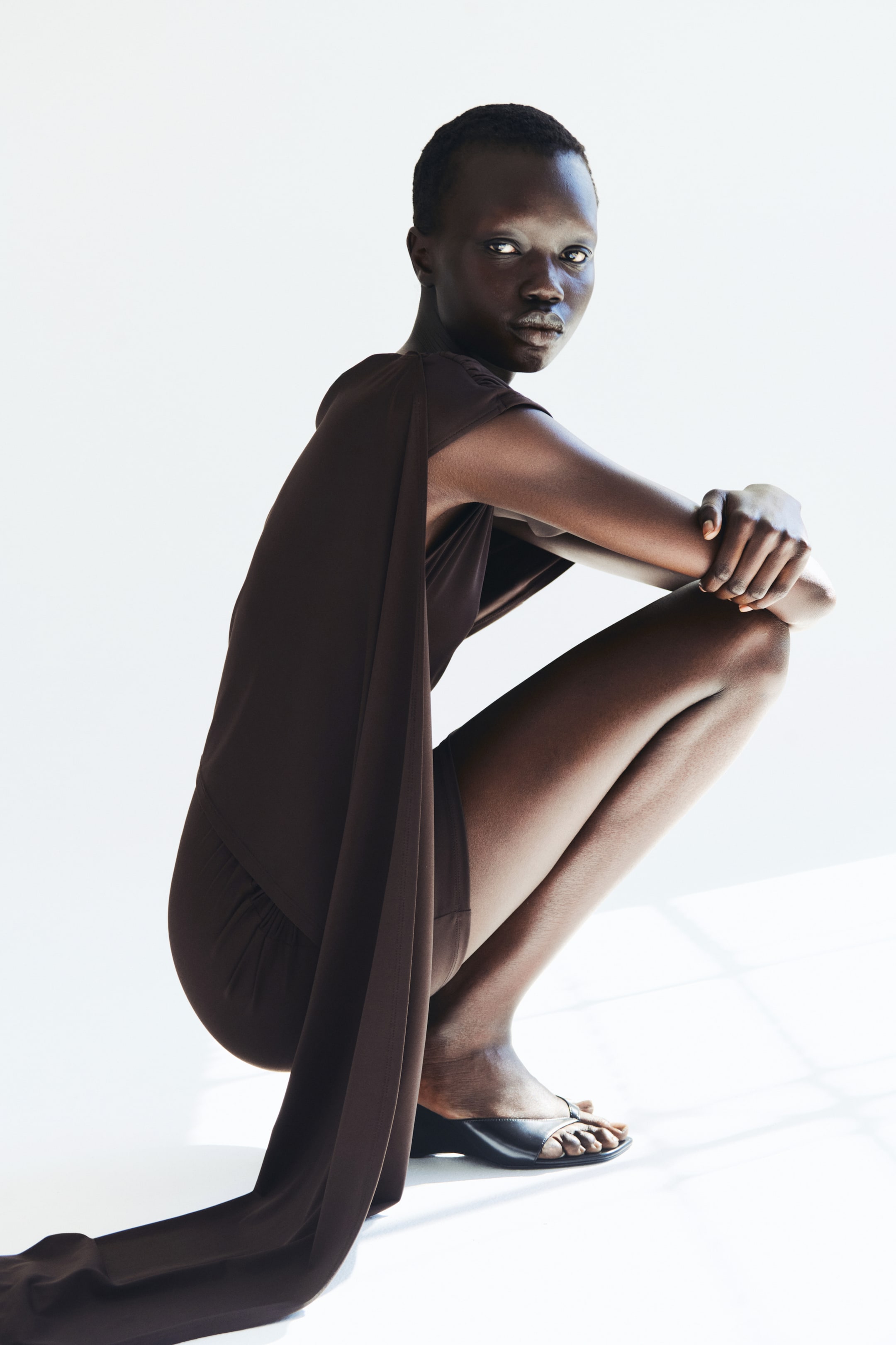 View larger image: The model is showcasing a fitted brown mini dress crafted from stretch jersey. This piece features gathered seams that deliver a draped effect, complemented by an asymmetric cape layer with an extended panel. The model is posed in a crouching position, paired with black wedge sandals.