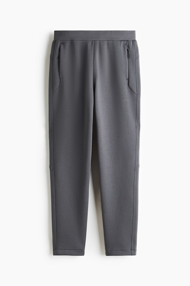 A pair of dark grey joggers is shown from the front, featuring an elastic waistband, vertical zippered side pockets, and tapered legs. The trousers are made from a smooth, structured fabric with seam detailing near the knees and neatly finished hems. - 1