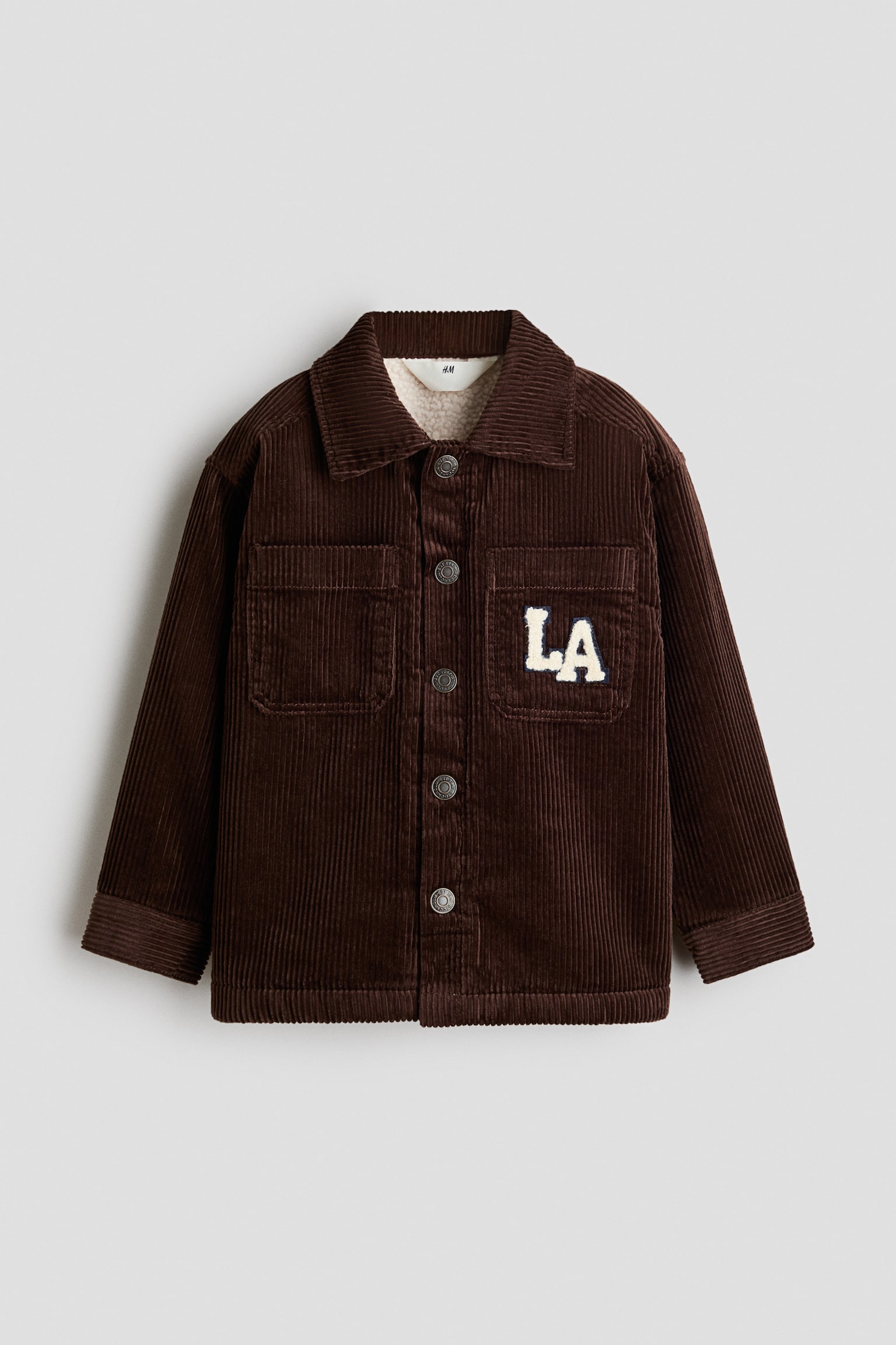 A dark brown corduroy jacket is shown from the front, featuring a classic collar, a button-up front with silver-tone metal buttons, and a visible beige sherpa lining at the neckline. The jacket is designed with two chest patch pockets, with a white varsity-style "LA" patch detailed on the left pocket.