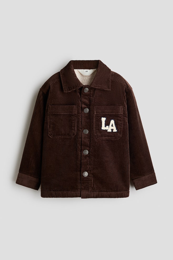 A dark brown corduroy jacket is shown from the front, featuring a classic collar, a button-up front with silver-tone metal buttons, and a visible beige sherpa lining at the neckline. The jacket is designed with two chest patch pockets, with a white varsity-style "LA" patch detailed on the left pocket. - 1