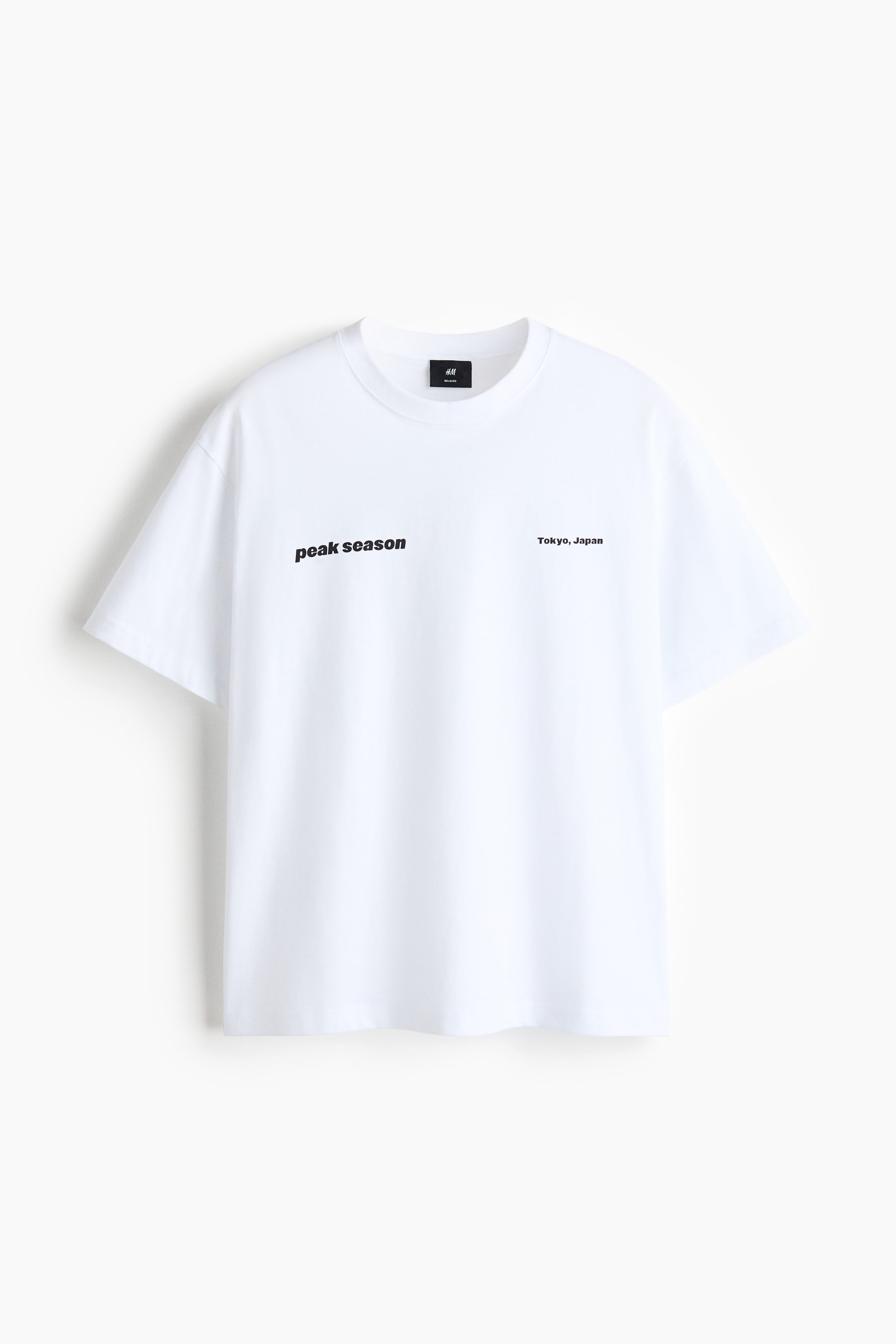 Grotere afbeelding bekijken: Een wit T-shirt van middelzwaar katoenjersey toont de voorkant, met een ronde, geribbelde halslijn, verlaagde schouders en een losse pasvorm. Het toont zwarte tekst "peak season" diagonaal gedrukt op de linkerborst en "Tokyo, Japan" diagonaal gedrukt op de rechterborst.