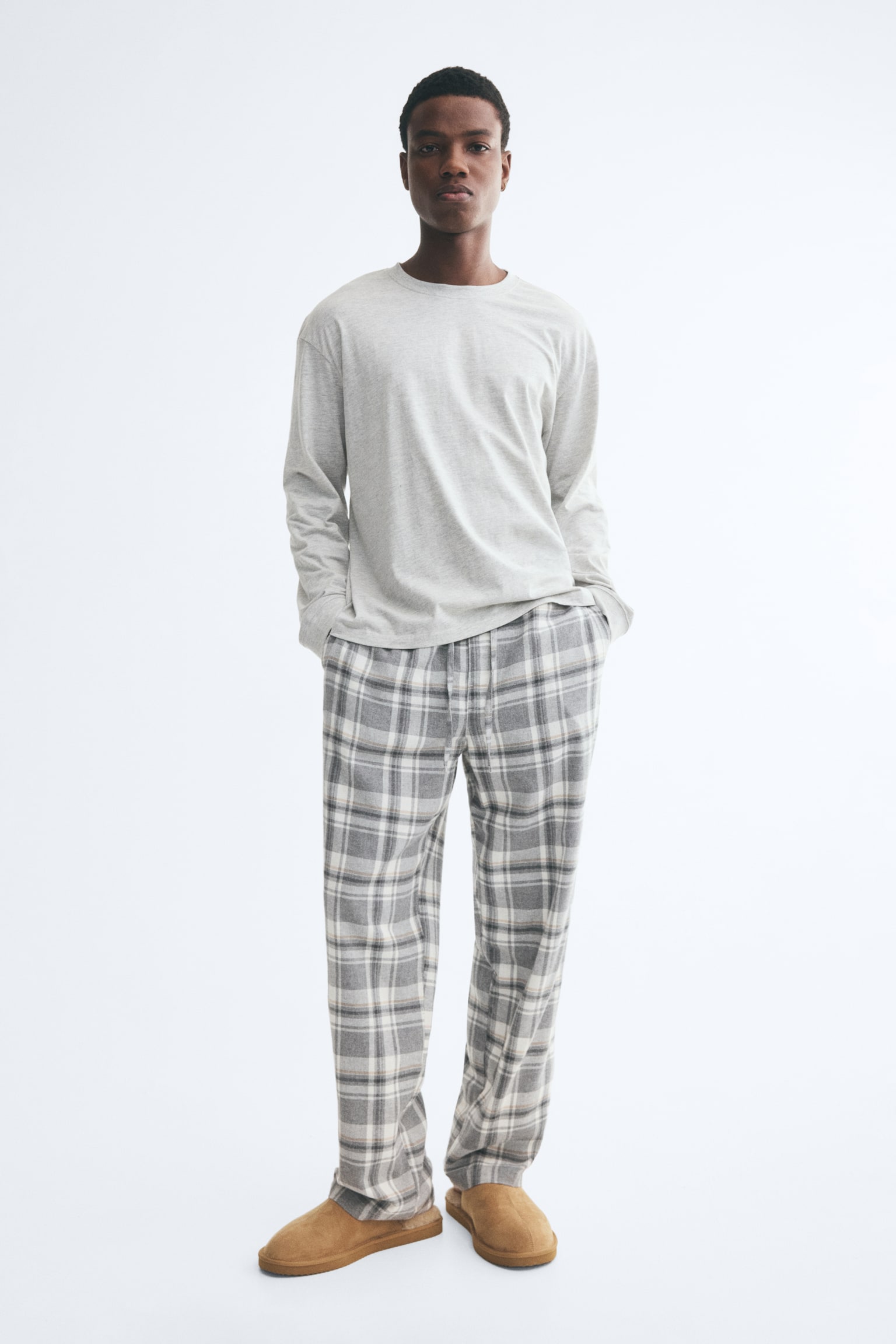 A pyjama set consisting of a light grey, long-sleeved jersey top and grey-and-white plaid flannel trousers is worn by the model. The model is standing with her hands placed inside the side pockets of the straight-leg trousers, which are paired with tan suede slippers. - 1