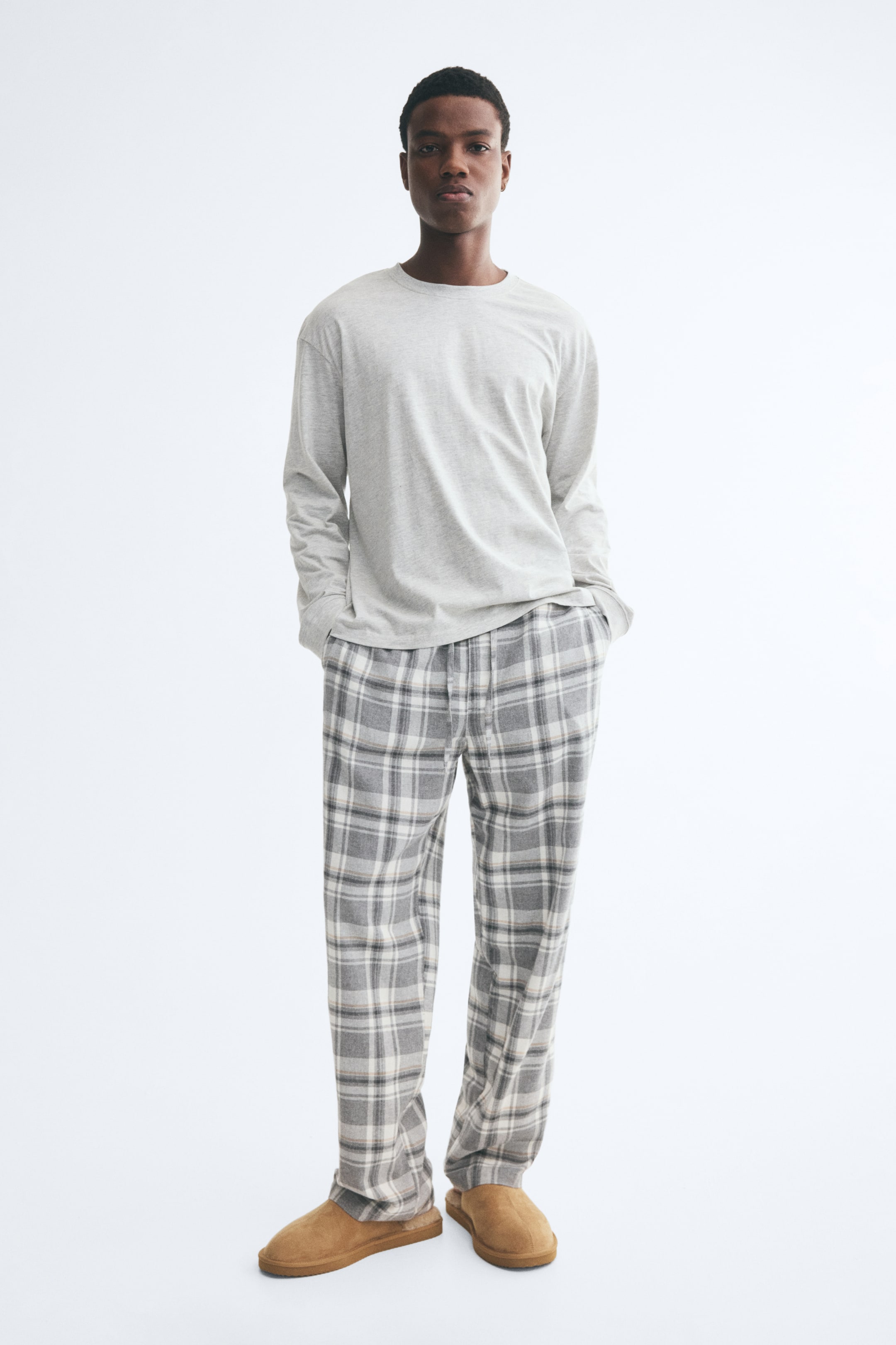 Visa större bild: Ett pyjamasset bestående av en ljusgrå, långärmad jerseytröja och rutiga flanellbyxor i grått och vitt bärs av modellen. Modellen står med händerna placerade i sidofickorna på de raka byxorna, som kombineras med tofflor i beige mocka.