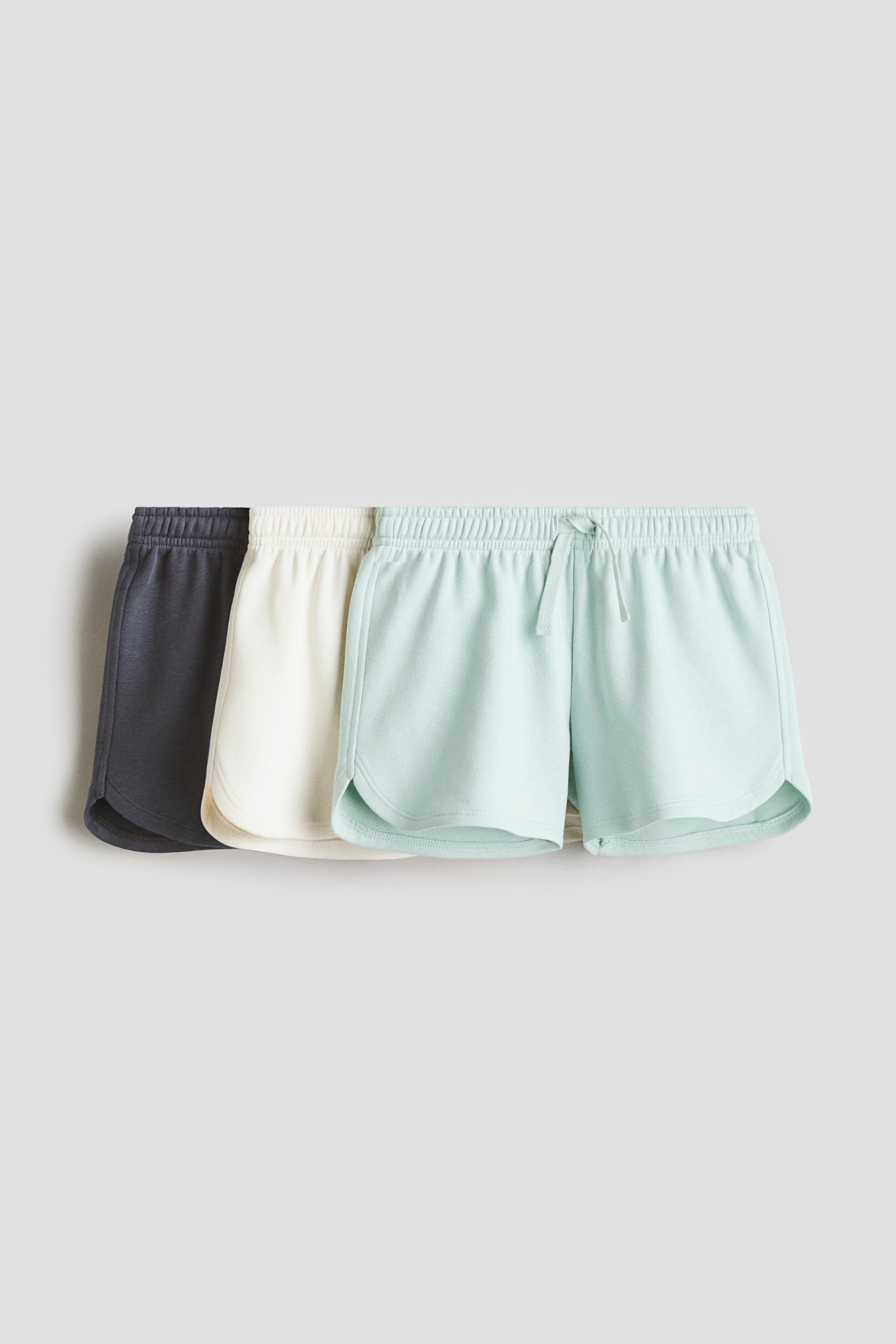 View larger image: Three pairs of jersey shorts are showcased in a layered arrangement, featuring dark grey, off-white, and light mint green shades, with the mint green pair prominently displayed at the front. Each short is equipped with an elasticated drawstring waistband and curved side hems, accented with matching piping.