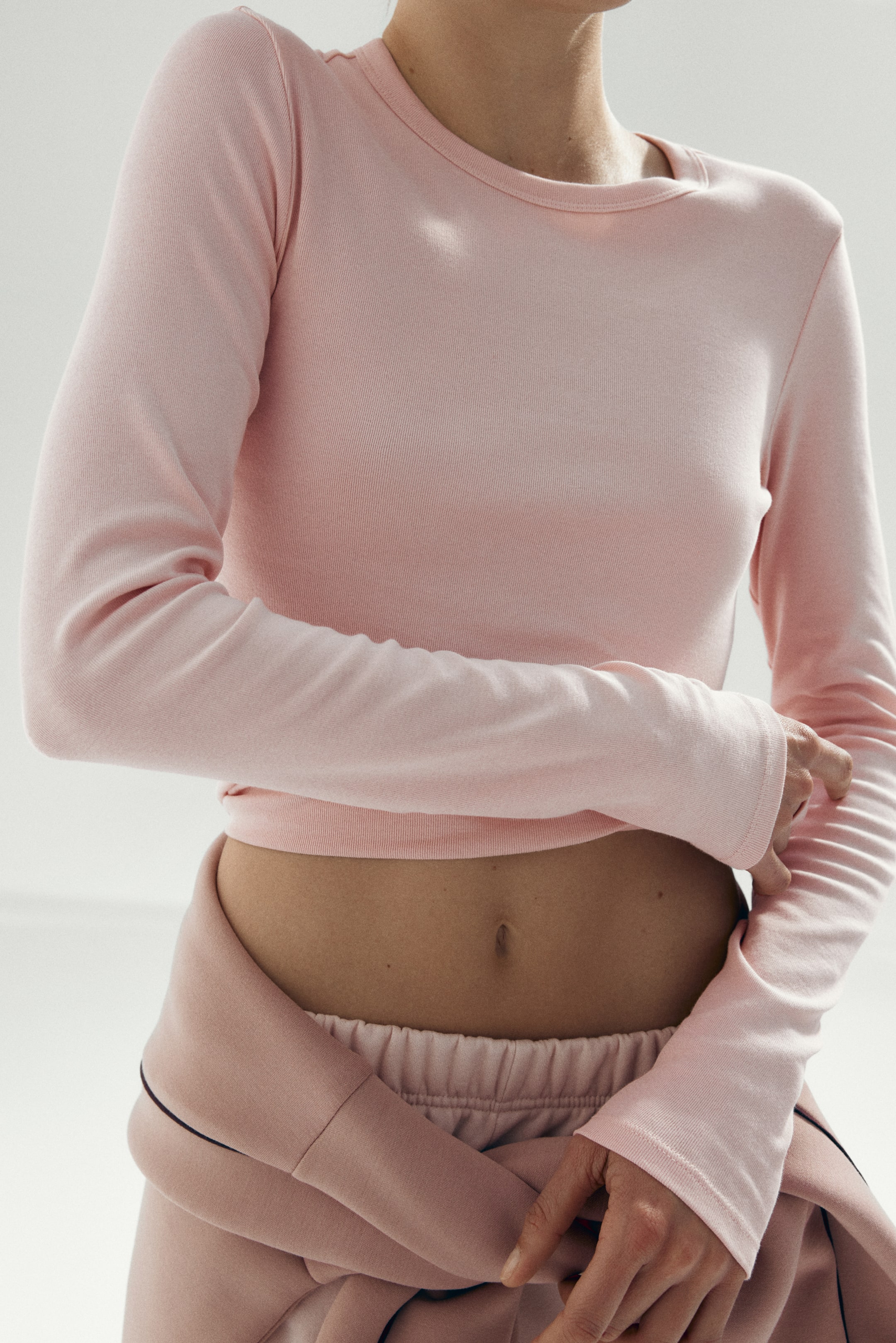 Agrandir l'image: Un haut court à manches longues rose est présenté de face, avec un col rond et une texture côtelée fine. Ses manches longues s'étendent jusqu'aux poignets, et l'ourlet court se termine au-dessus de la taille, où un pantalon de survêtement rose est visible.