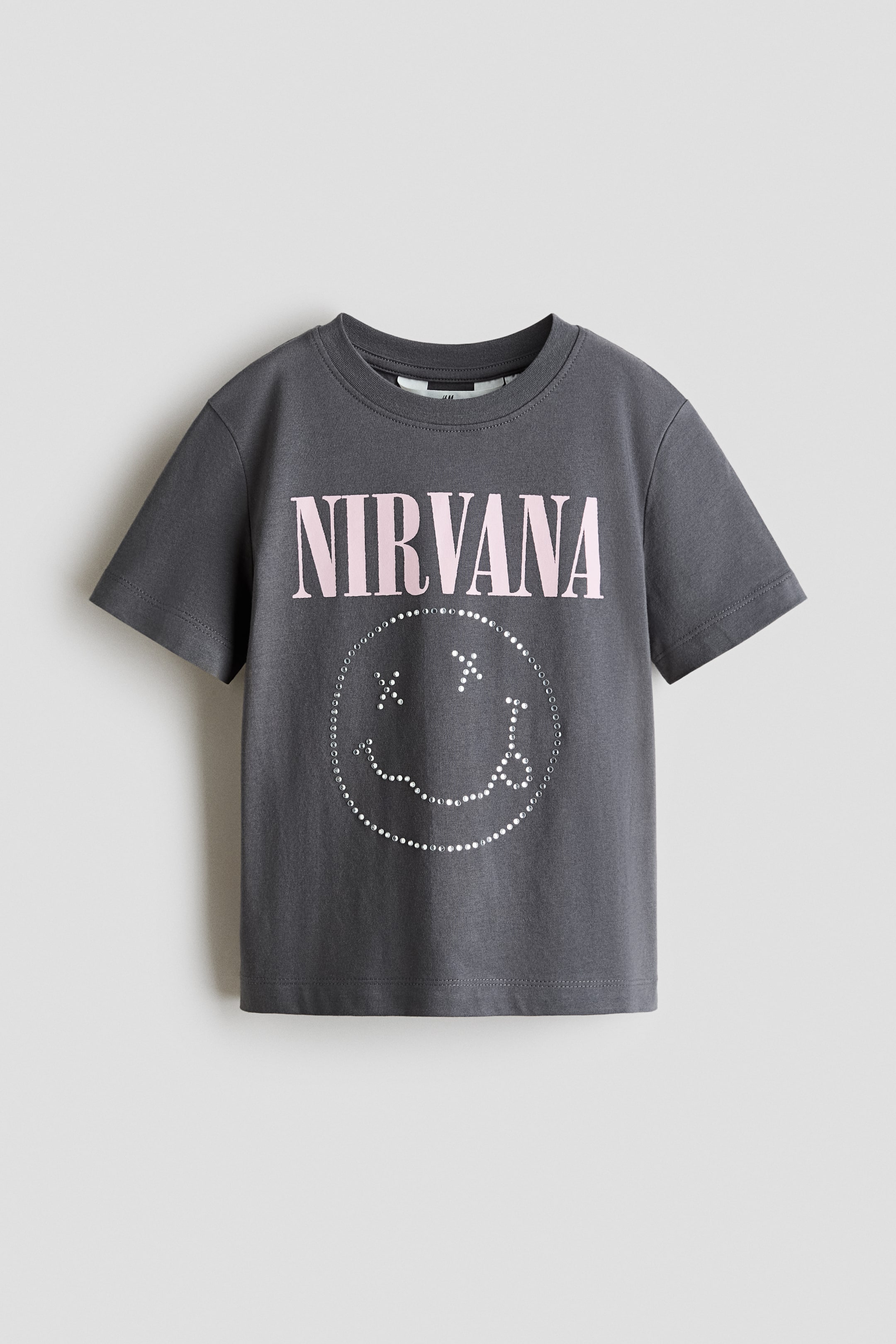 Grösseres Bild ansehen: Ein dunkelgraues T-Shirt aus weichem Baumwolljersey ist von vorne zu sehen, mit einem runden Halsausschnitt und kurzen Ärmeln. Auf der Vorderseite ist ein hellrosa "NIRVANA"-Druck zu sehen, darunter bildet sich ein Smiley-Gesicht, das mit glänzenden Nieten umrandet ist.