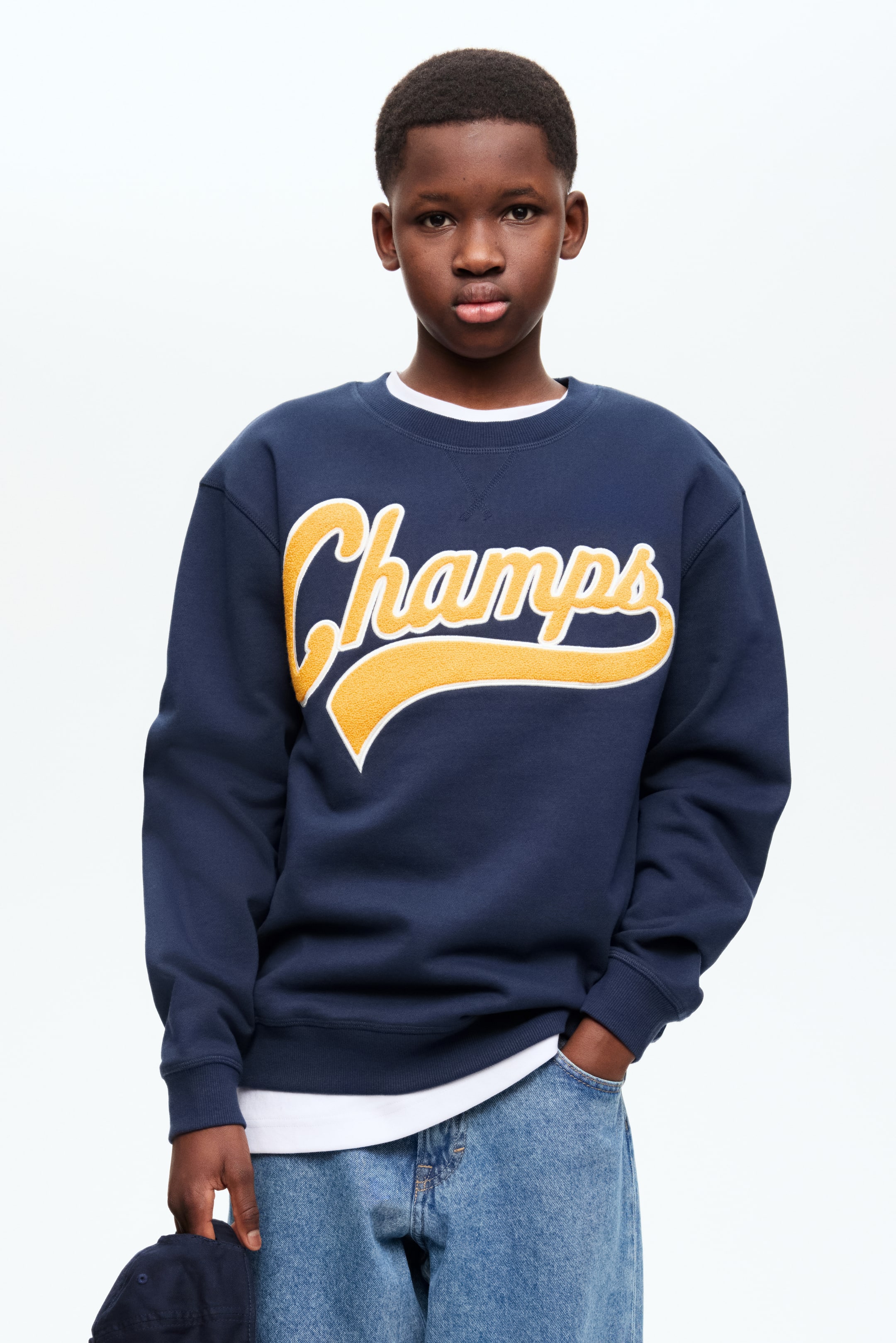 查看大圖: 這款海軍藍輕薄運動衫面料的上衣，正面印有黃色「Champs」圖案，並以白色輪廓勾勒。設計上採用圓形羅紋領口，落肩設計，袖口和下擺均有羅紋裝飾，並搭配一件白色T恤穿著。