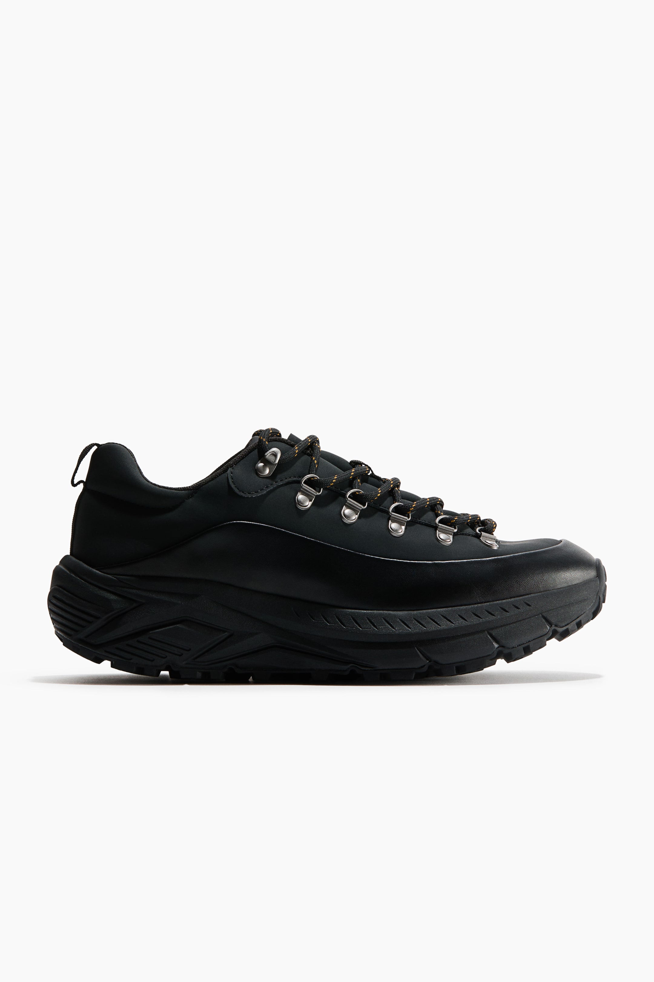 View larger image: A low-top black hiking shoe is presented in side profile, featuring a smooth matte upper with silver-tone D-ring eyelets holding black and orange speckled laces. The shoe showcases a thick, textured black midsole with a rugged, lugged outsole and a heel pull tab.