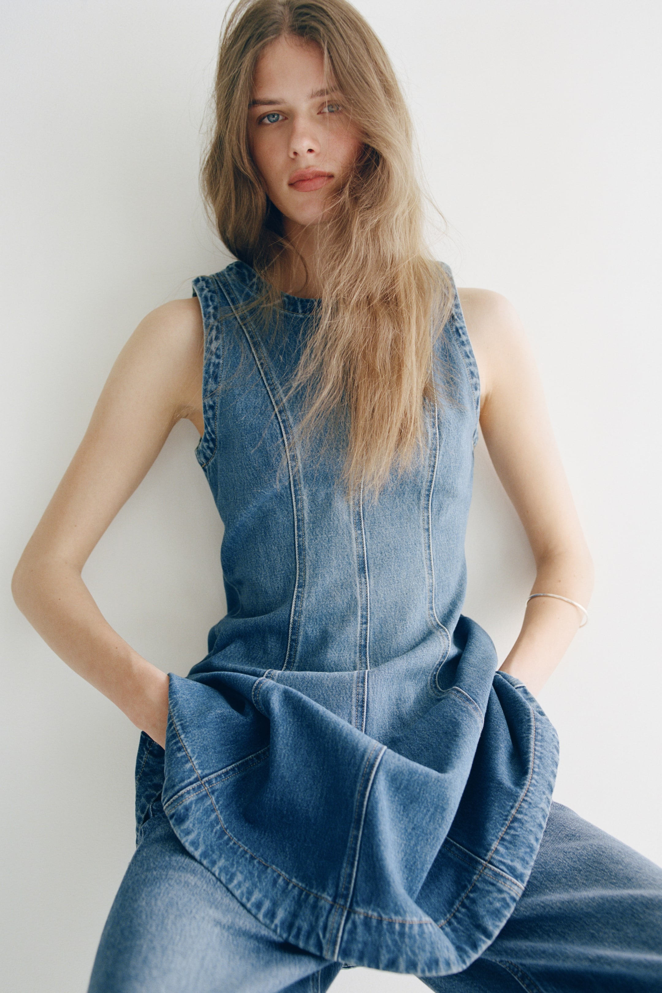 Größeres Bild anzeigen: Der blaue Denim-Jumpsuit wird leicht nach vorne gerichtet präsentiert und zeigt ein ärmelloses Oberteil mit vertikalen Nahtdetails sowie weite Hosenbeine, wobei die Hände in den sichtbaren Seitentaschen ruhen.