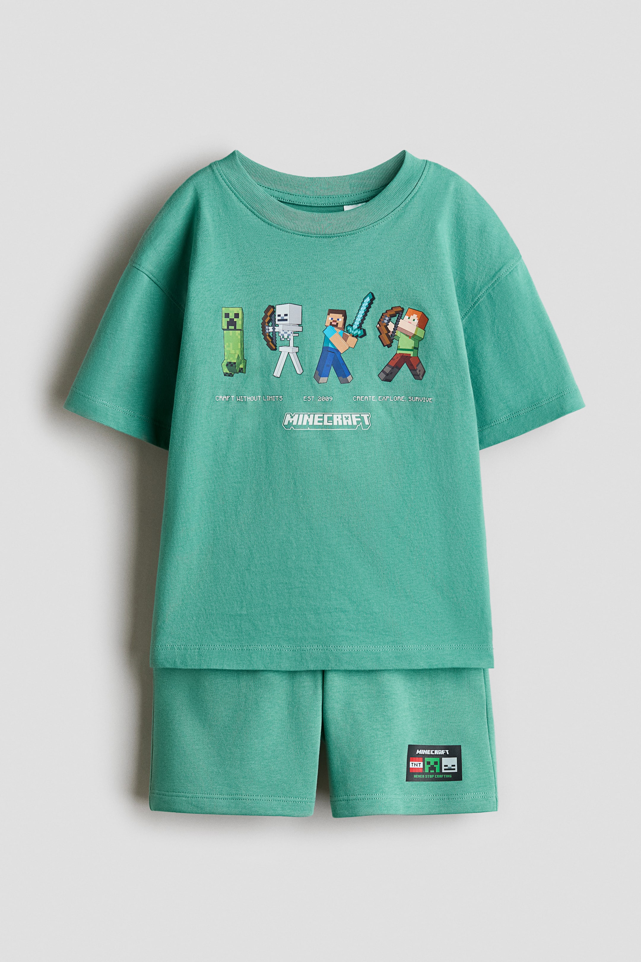 View larger image: A matching teal set features a T-shirt and shorts. The T-shirt, shown front-facing, displays Minecraft characters and the logo on the front, with dropped shoulders, wide sleeves, and a ribbed neckline. The shorts have a covered elastic waistband and a small "Minecraft" label on the left leg.