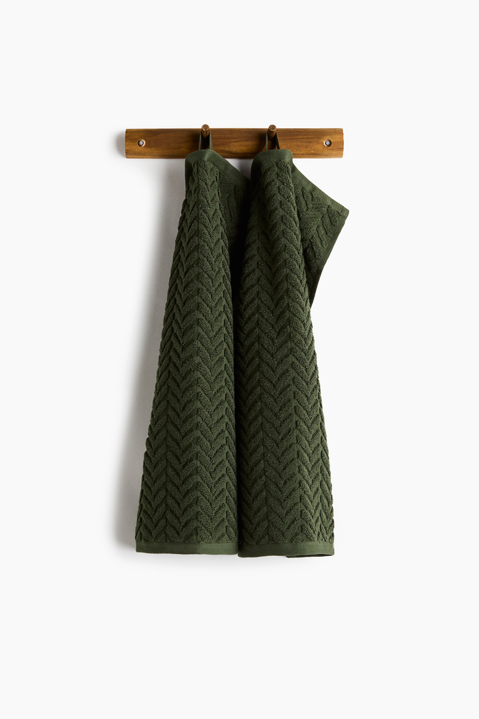 A pair of dark green cotton terry hand towels is hanging side-by-side from a wooden peg rack, displaying a textured, cable-knit-inspired chevron pattern. Each towel is suspended by a loop on its short side, showcasing the soft, draped fabric.