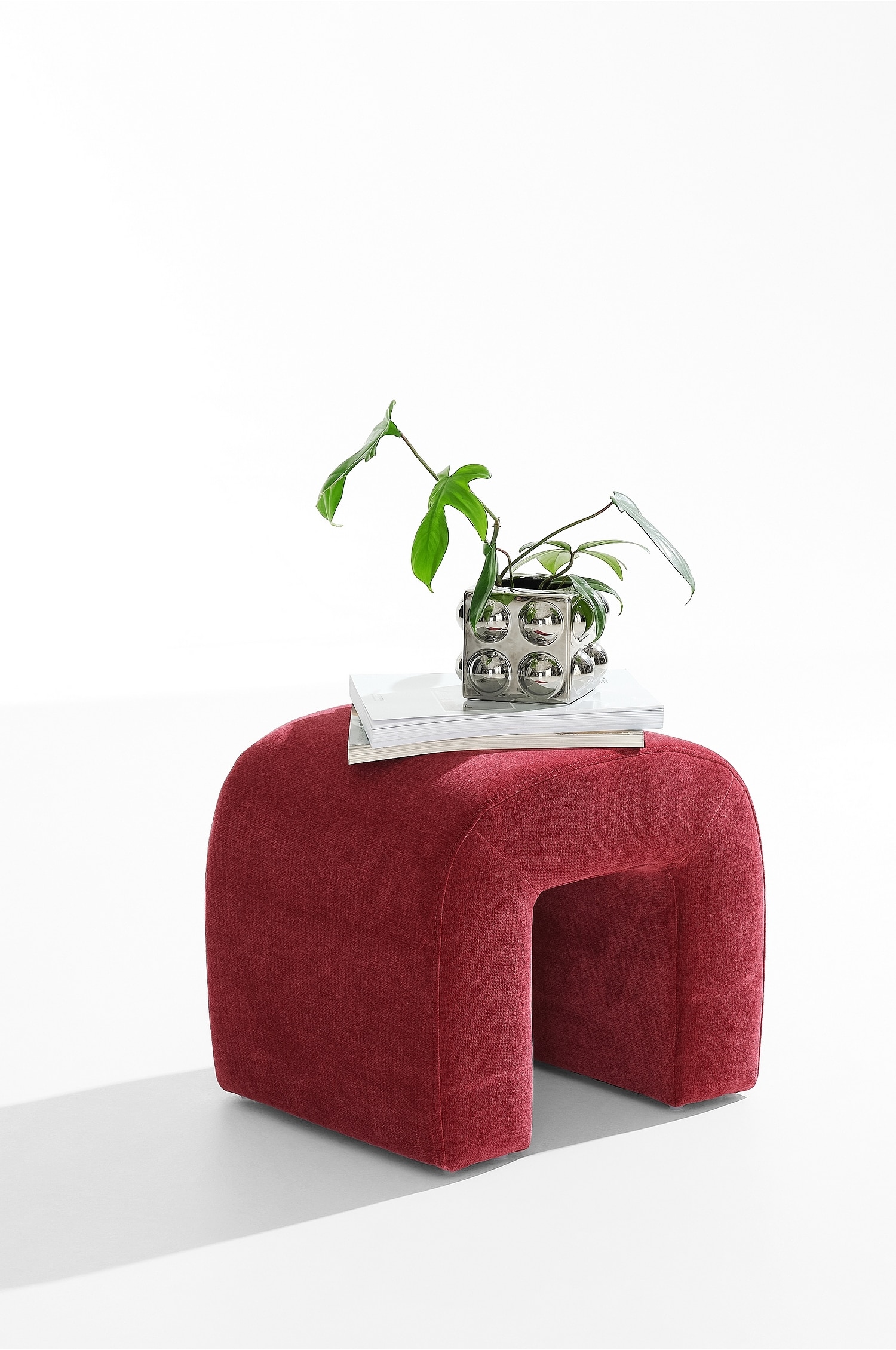 Un pouf in velluto rosso dalla forma a U rovesciata è posizionato in una vista a tre quarti, mettendo in risalto la seduta arrotondata e imbottita e lâarco cavo. Lo sgabello è rivestito in velluto testurizzato color cremisi con sottili cuciture in evidenza.