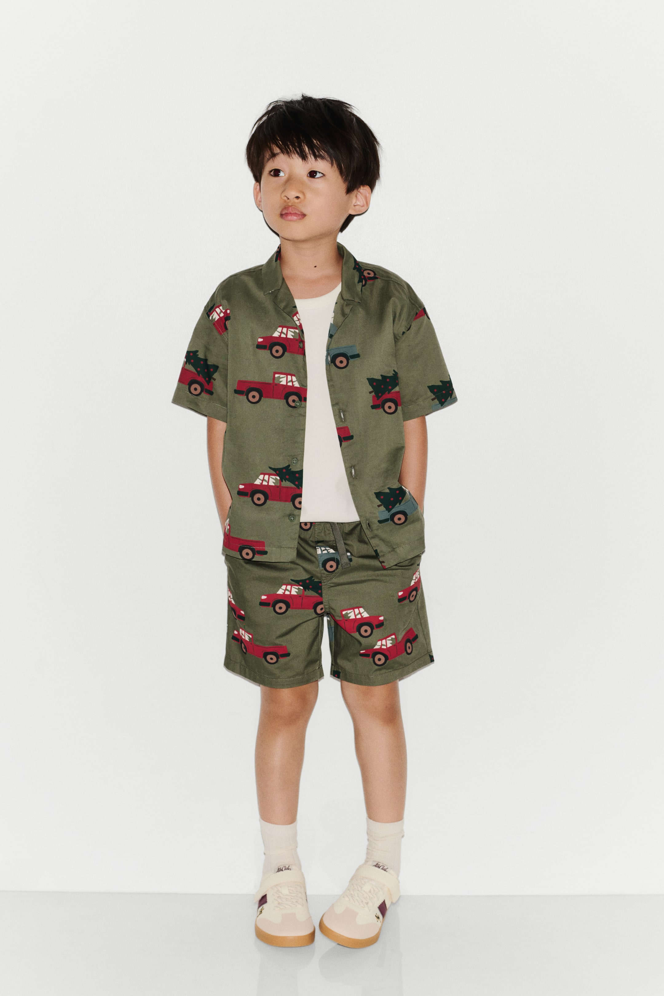 View larger image: These olive green cotton-weave shorts, featuring an all-over print of red and blue trucks carrying pine trees, are shown from the front on a model. The shorts feature an elasticated waistband with a drawstring, diagonal side pockets, and a mock fly, and are styled with a matching short-sleeve shirt.
