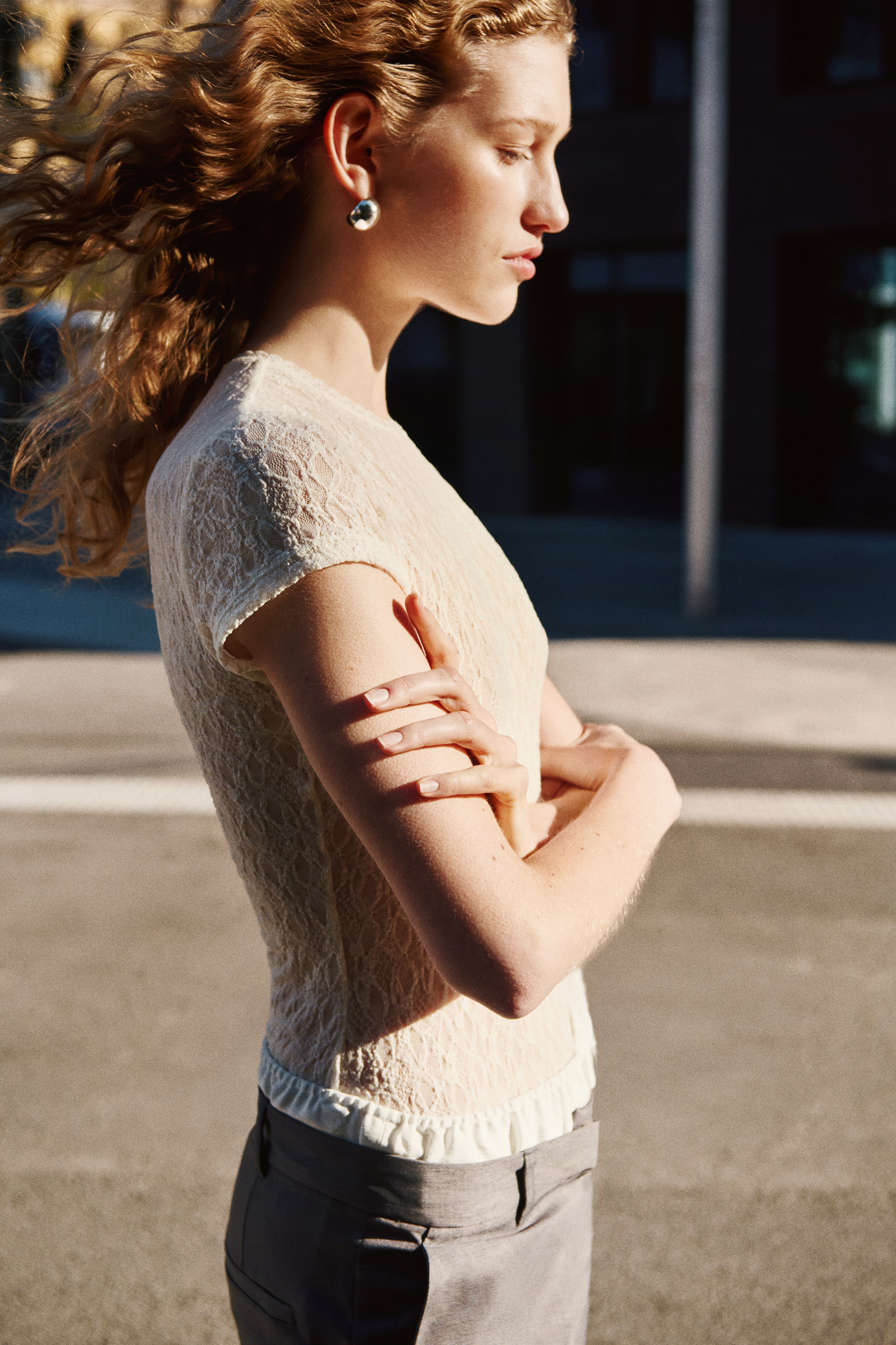 View larger image: The cream lace top showcases a tailored silhouette from a side perspective, highlighted by short sleeves and an intricate lace pattern. Its ruffled hem gracefully sits above grey trousers.