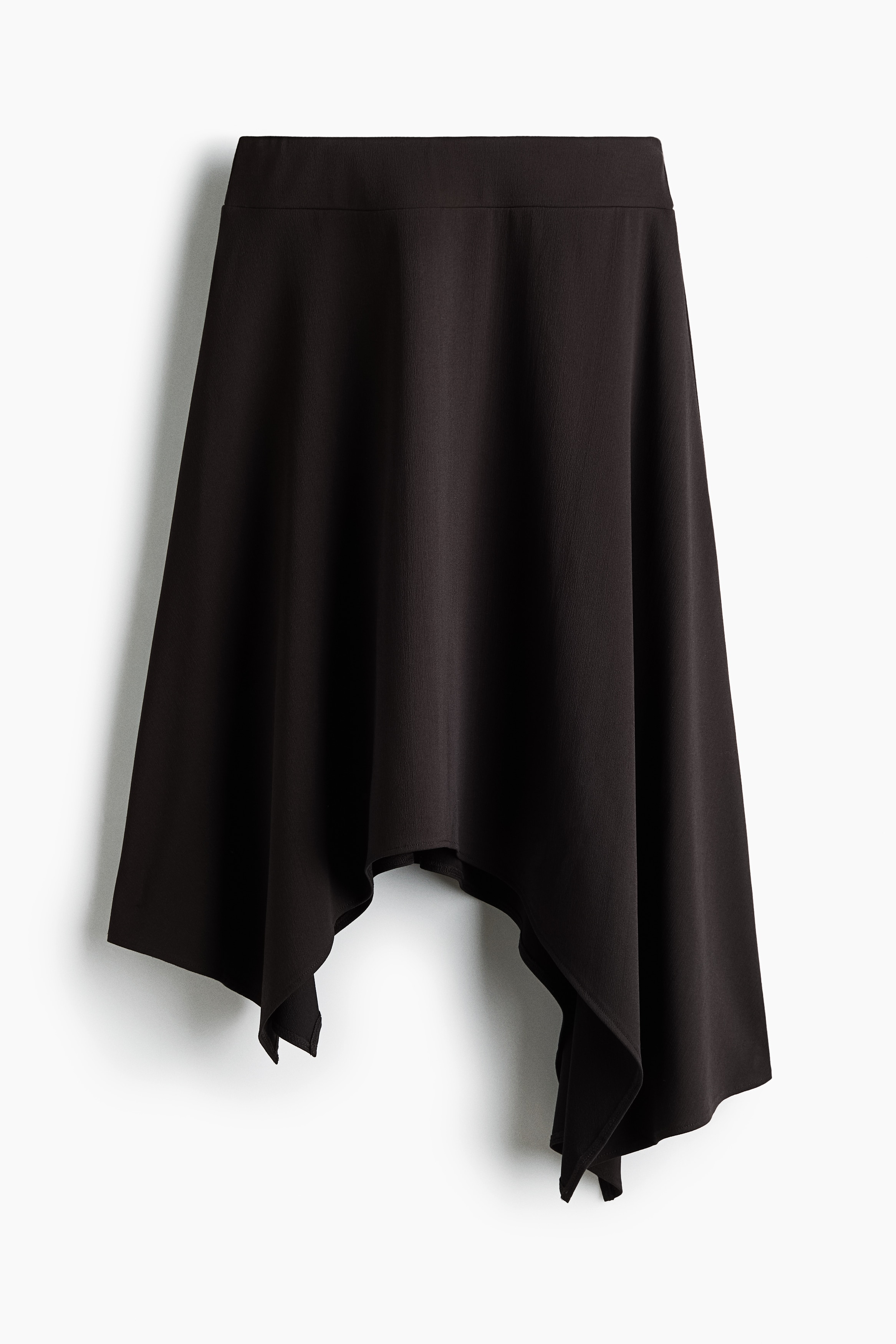 A dark brown, calf-length skirt in textured stretch jersey is worn by the model, featuring a loose A-line fit and an asymmetrical handkerchief hem. The skirt is styled with a high-collared black leather jacket layered over the waist.