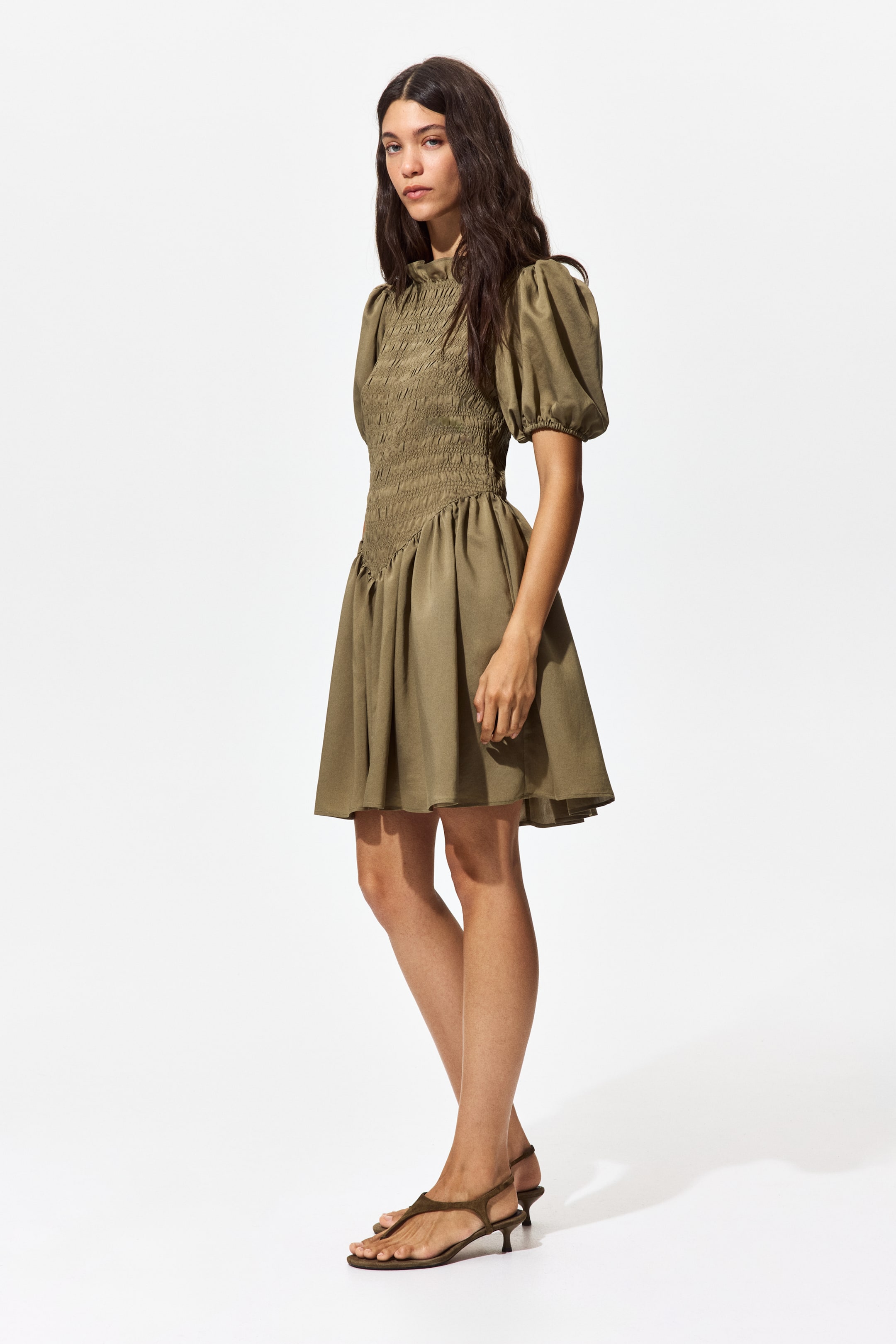 Ampliar la imagen: Un vestido mini verde oliva presenta un cuello alto y mangas cortas voluminosas. El vestido está diseñado con un corpiño fruncido que tiene un borde inferior en forma de V, fluyendo hacia una falda en línea A.