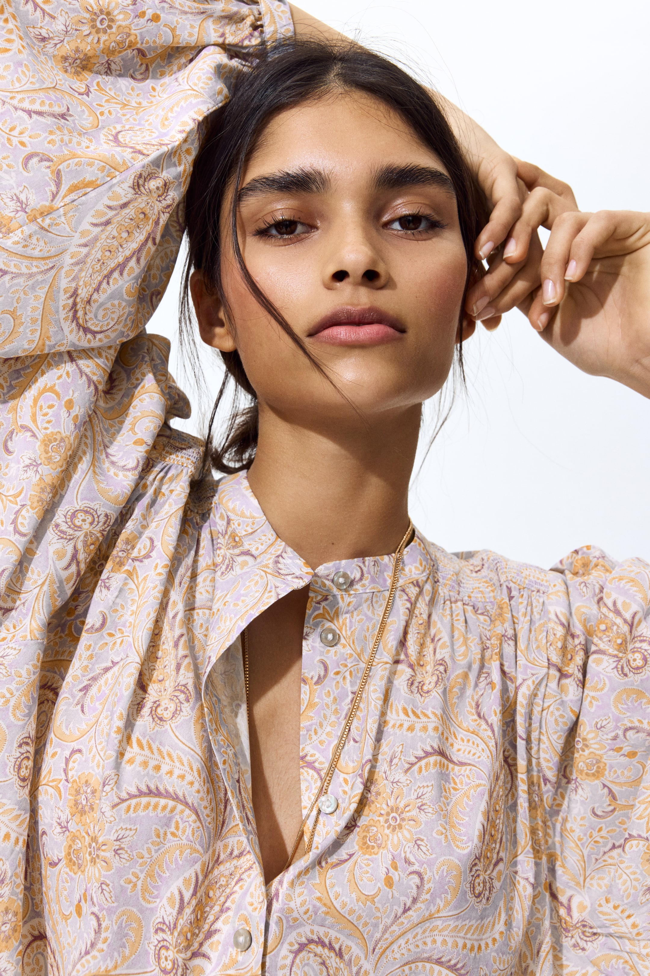 View larger image: An oversized blouse, showcasing a paisley pattern in purple and yellow hues, is being worn. It comes with a band collar, pearl-like buttons along the front, and smocked detailing on the shoulders with balloon sleeves. The model is raising her hands, and a delicate gold chain necklace is resting over the partially unbuttoned front of the blouse.
