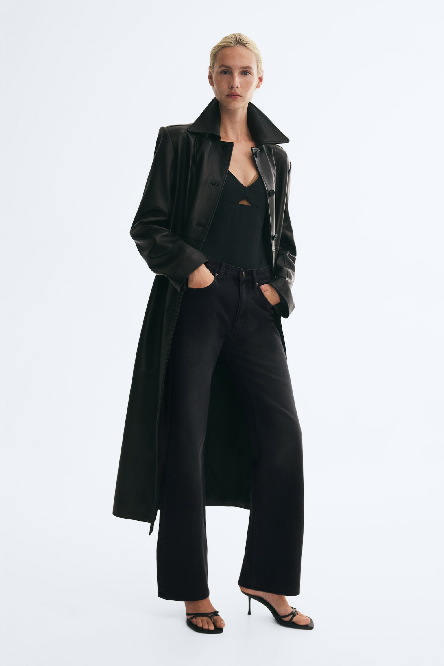 Schwarze High-Waist-Denim-Jeans werden von dem Model getragen, das mit den HÃ¤nden in den vorderen Taschen steht. Die Jeans sitzen an Taille und Oberschenkeln schmal und laufen am Saum leicht aus. Dazu werden ein schwarzes Oberteil, ein langer schwarzer Ledermantel und schwarze Sandaletten mit Absatz kombiniert. - 1