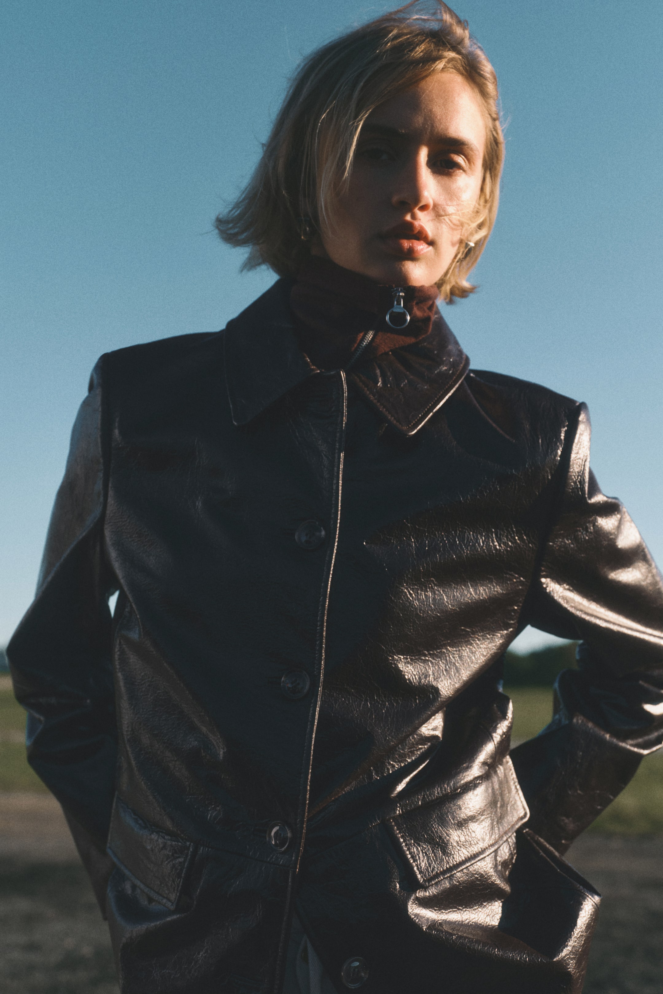 Ingrandisci l'immagine: Una giacca scura e lucida è indossata, caratterizzata da un colletto classico, una chiusura frontale con bottoni e due tasche con patta inferiori. Un dolcevita rosso scuro con collo con cerniera e anello è sovrapposto sotto.