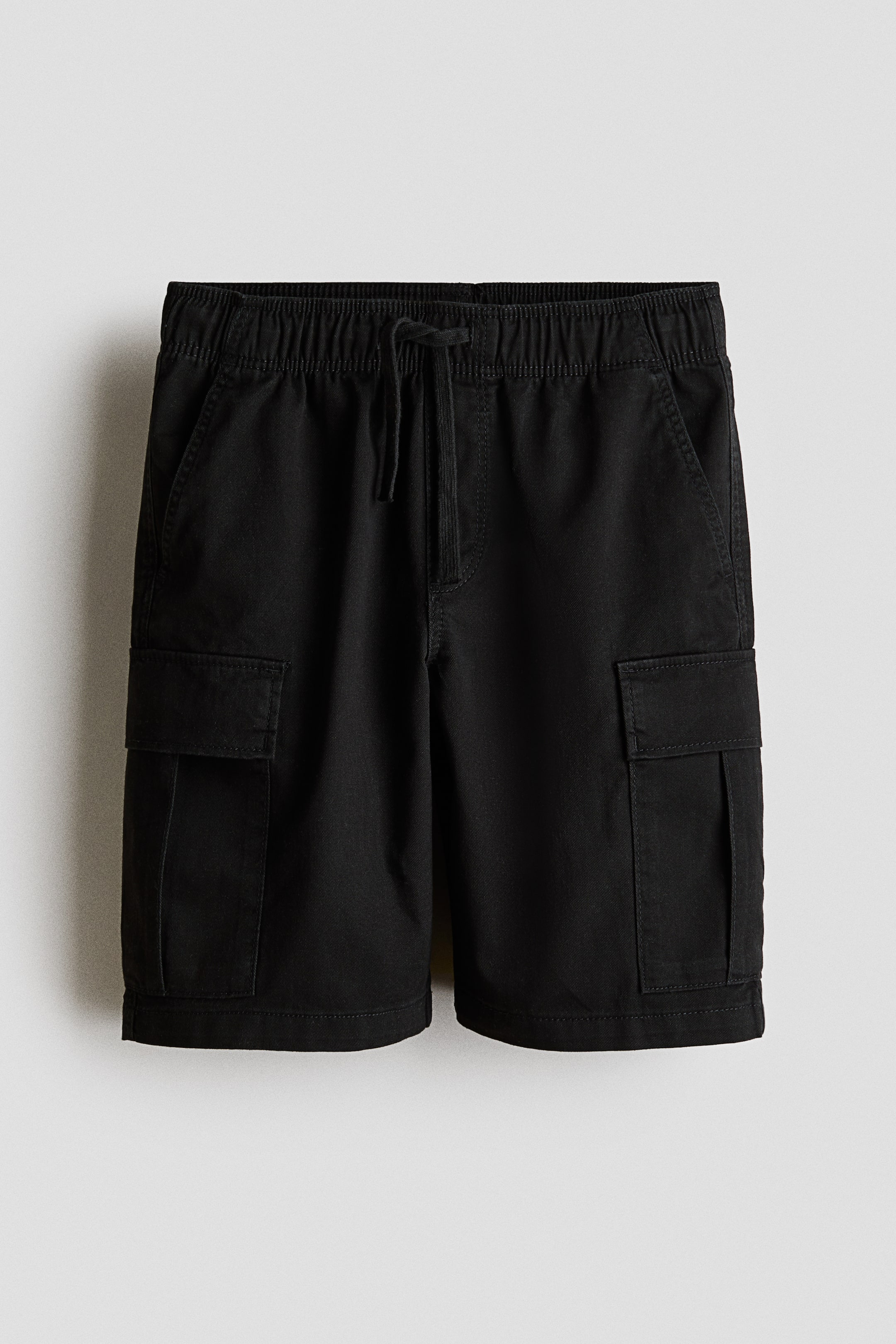 View larger image: Black knee-length shorts are shown from the front. They feature an elasticated drawstring waist, a mock fly, diagonal side pockets, and two flap leg pockets.