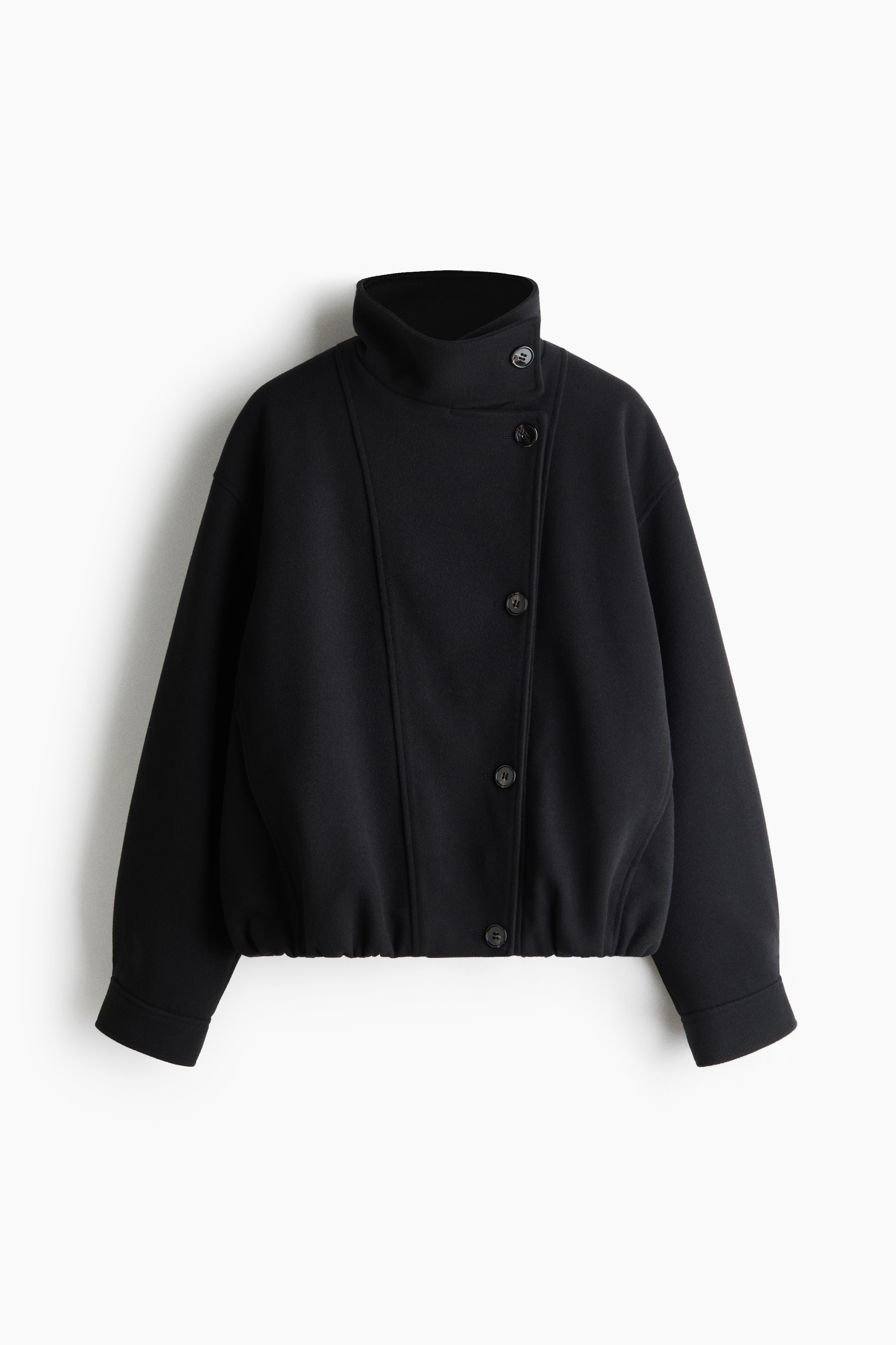 Une veste noire oversize en tissu feutrÃ© doux est portÃ©e par le mannequin, avec un col cheminÃ©e boutonnÃ© et une fermeture Ã  boutons dÃ©calÃ©e sur le devant. Le mannequin se tient debout, les mains glissÃ©es dans les poches dâun jean en denim bleu visible sous lâourlet de la veste.
