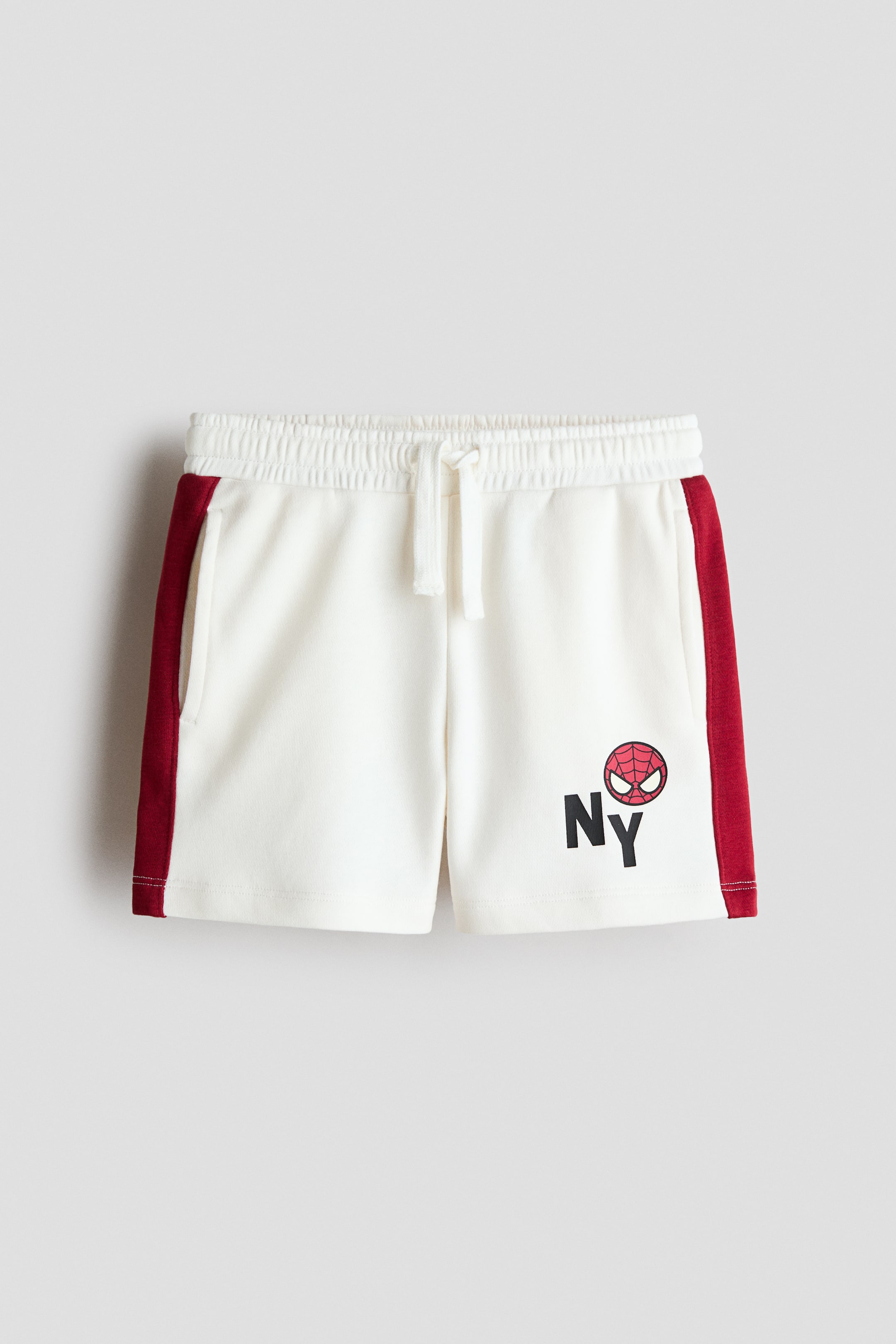 View larger image: Off-white shorts are shown from the front, showcasing an elasticated waistband with a white drawstring and dark red side stripes running down each leg. An inseam pocket is visible on the left, and an "NY" graphic print with a red and black Spider-Man face replacing the "Y" appears on the lower right leg.