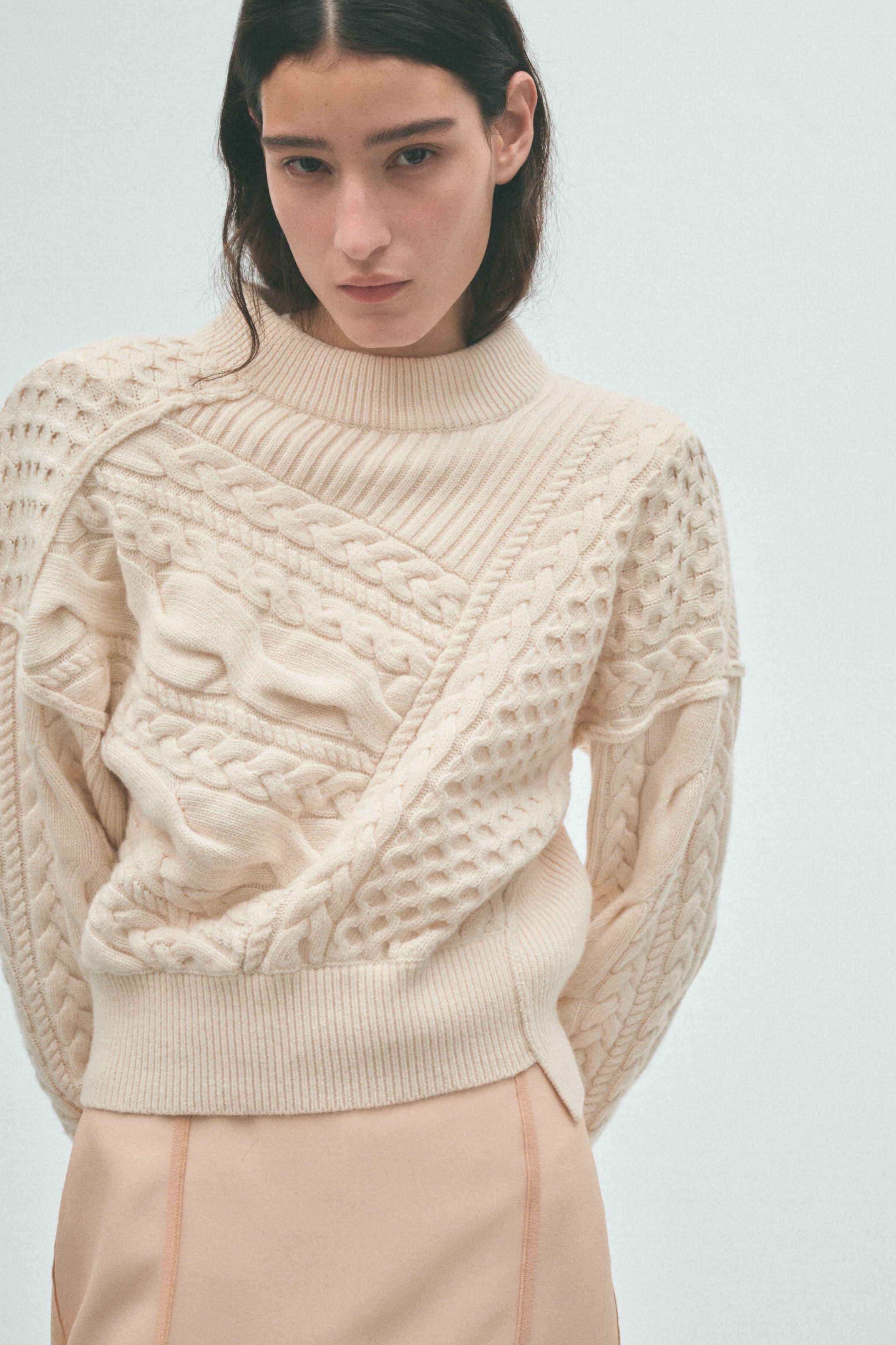View larger image: A cream knitted jumper is being worn by the model, featuring a high neck and various cable knit and textured patterns. An asymmetrical design crosses the front, creating a diagonal textured panel.