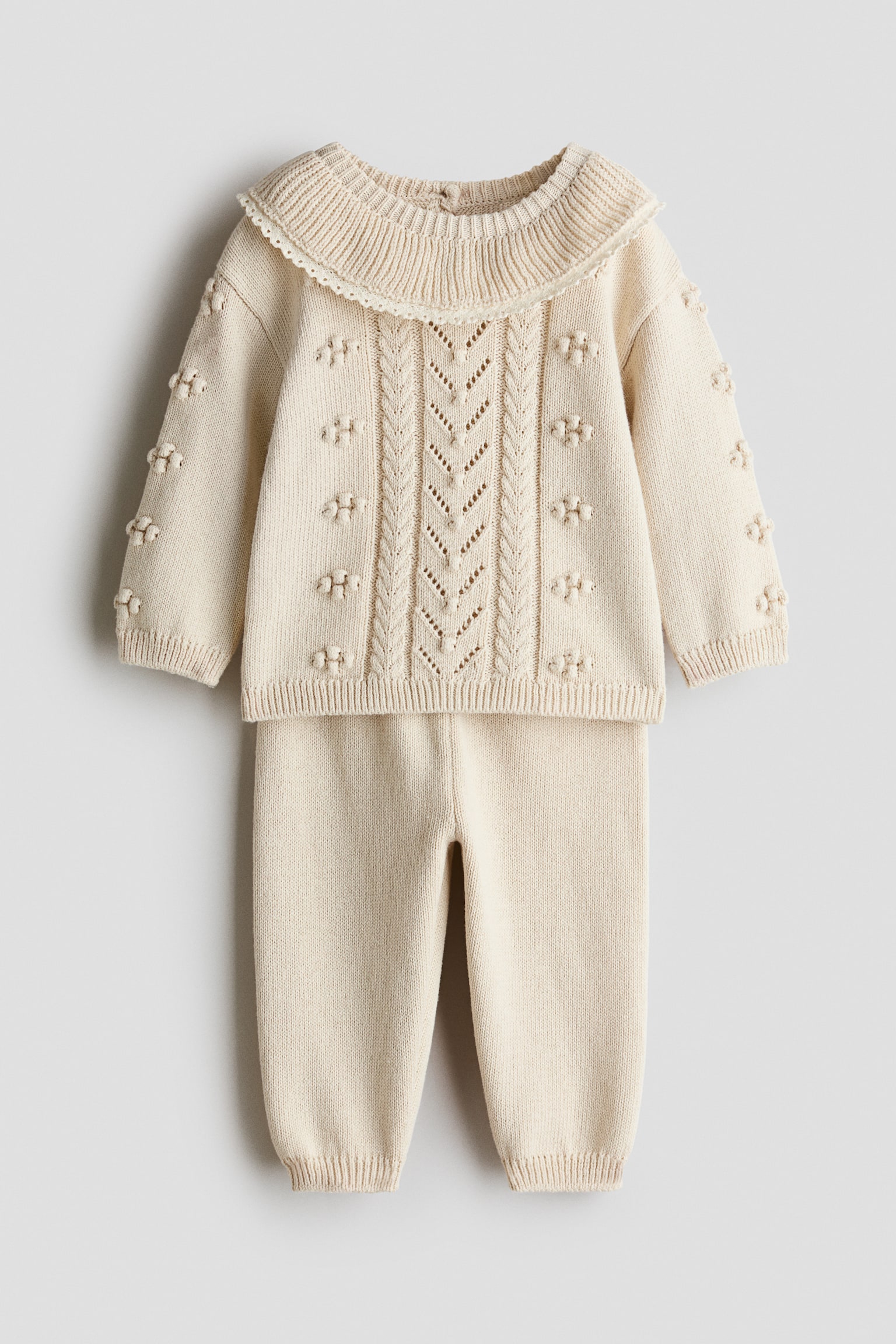 Ett babyset i krÃ¤mfÃ¤rgad stickad kvalitet visas i framifrÃ¥n med en lÃ¥ngÃ¤rmad trÃ¶ja och matchande byxor. TrÃ¶jan har en bred ribbad krage med vÃ¥gad spetskant, centrala kabelstickade och pointelle-mÃ¶nster samt upphÃ¶jda stickade noppor pÃ¥ framsidan och Ã¤rmarna. De matchande byxorna gÃ¥r ner med en ren stickad design och ribbade fotleder med muddar. - 1