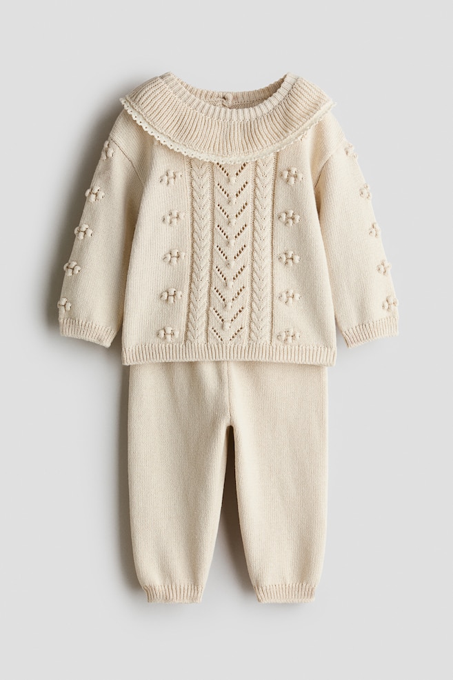 A cream-coloured knit baby set is displayed in a front view, featuring a long-sleeved jumper and matching trousers. The jumper features a wide, ribbed collar with a scalloped lace edge, central cable-knit and pointelle patterns, and raised knit bobbles on the front and sleeves. The matching trousers extend down with a clean knit design and ribbed ankle cuffs. - 1