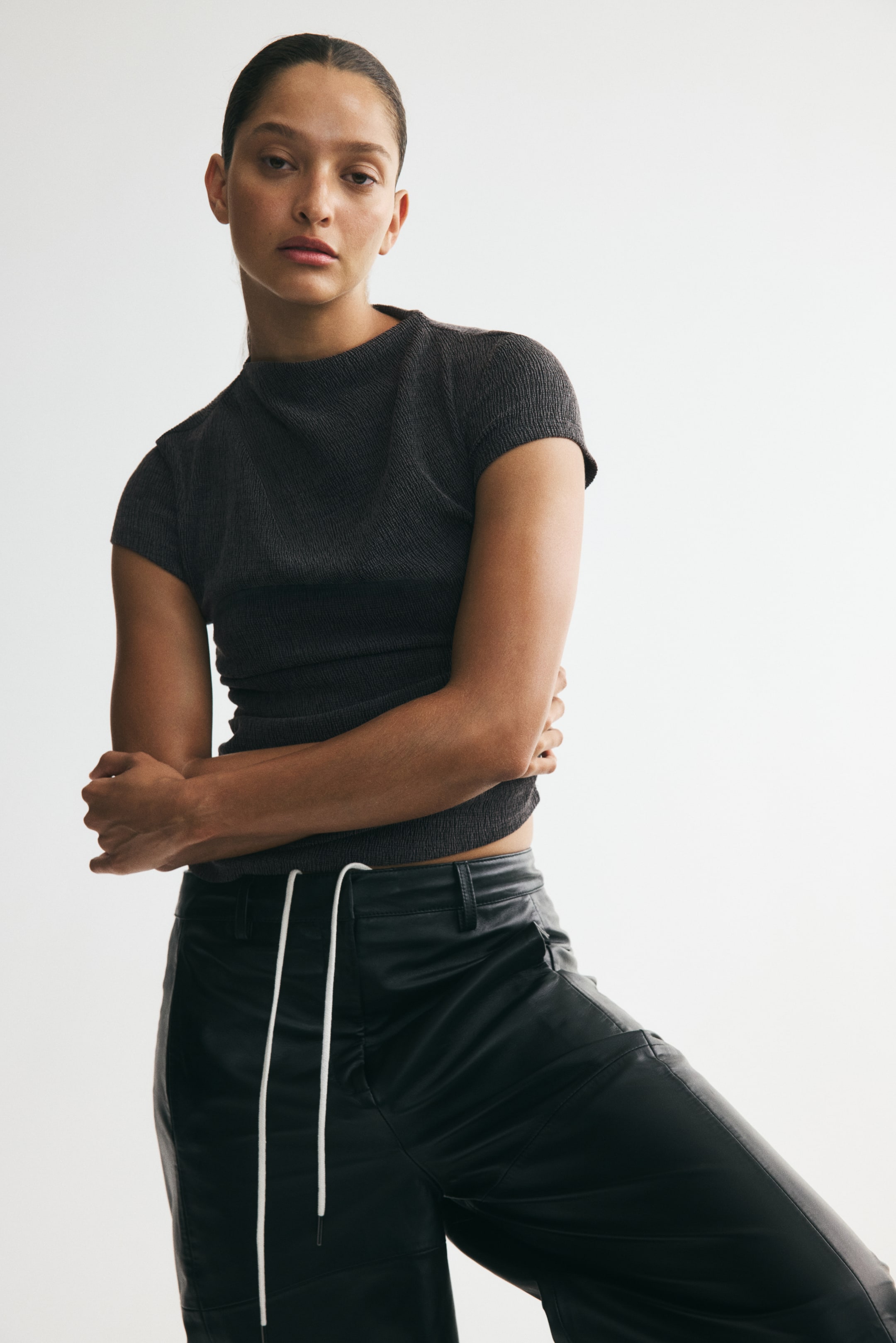 View larger image: The model is wearing a dark grey fitted top crafted from crinkled jersey, featuring a stand collar and cap sleeves. The model is posed with their arms crossed in front of the top.