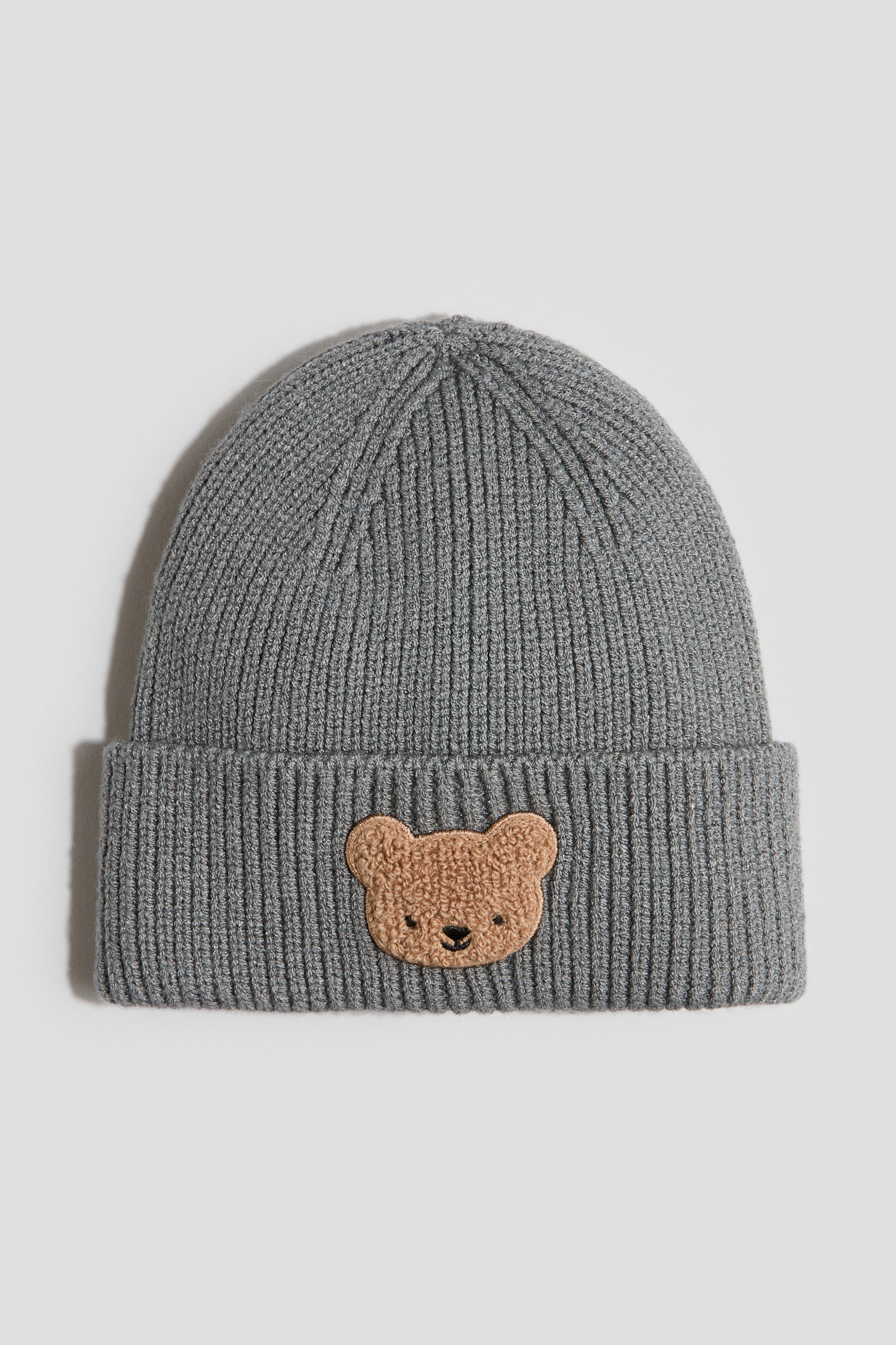 View larger image: A grey rib-knit beanie is shown facing forward, featuring a brown teddy bear face appliqué on its folded cuff.