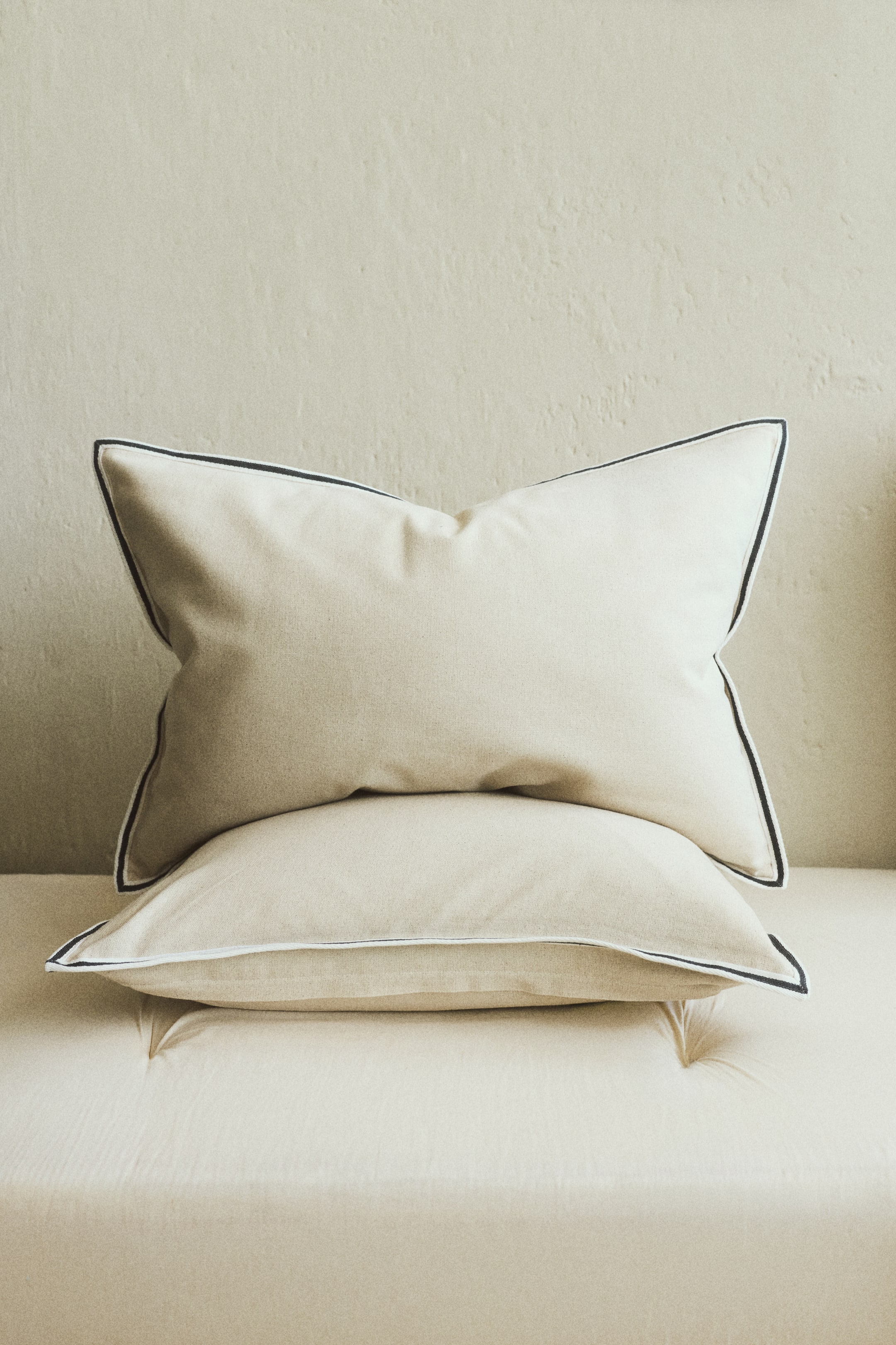 Voir une image plus grande: Deux coussins beige clair, fabriqués dans un tissu doux et brossé, sont posés l'un sur l'autre. Les deux coussins présentent un passepoil fin blanc et foncé le long de leurs bords, avec le coussin supérieur incliné vers l'avant et à droite.