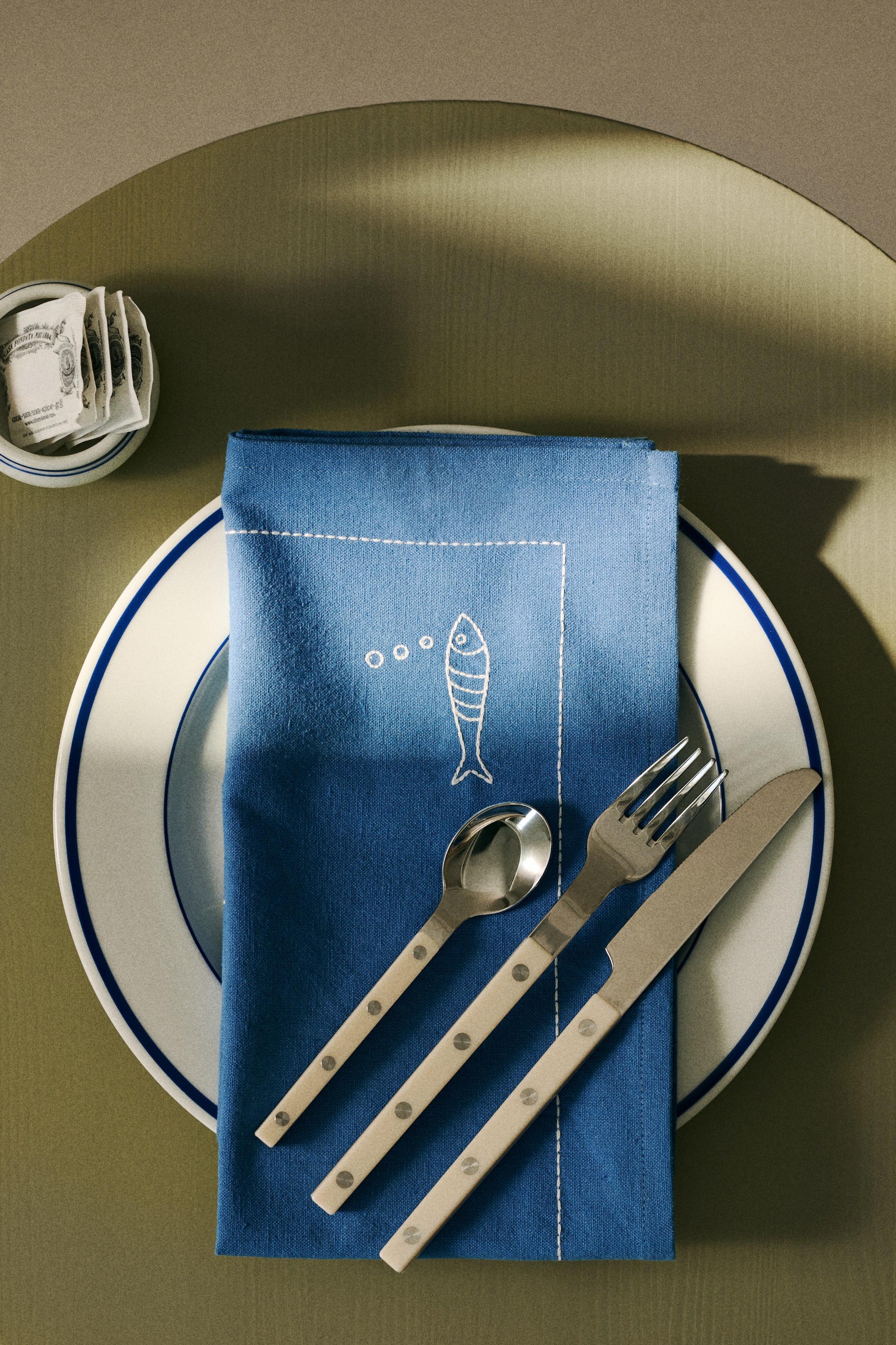 Zobacz większy obraz: Ręcznik kuchenny z mieszanki bawełny i lnu leży złożony na białym talerzu, z sztućcami spoczywającymi na nim. Charakteryzuje się niebieskim odcieniem z białym haftem w postaci przerywanej linii tworzącej obramowanie oraz białym haftowanym wzorem ryby z bąbelkami.