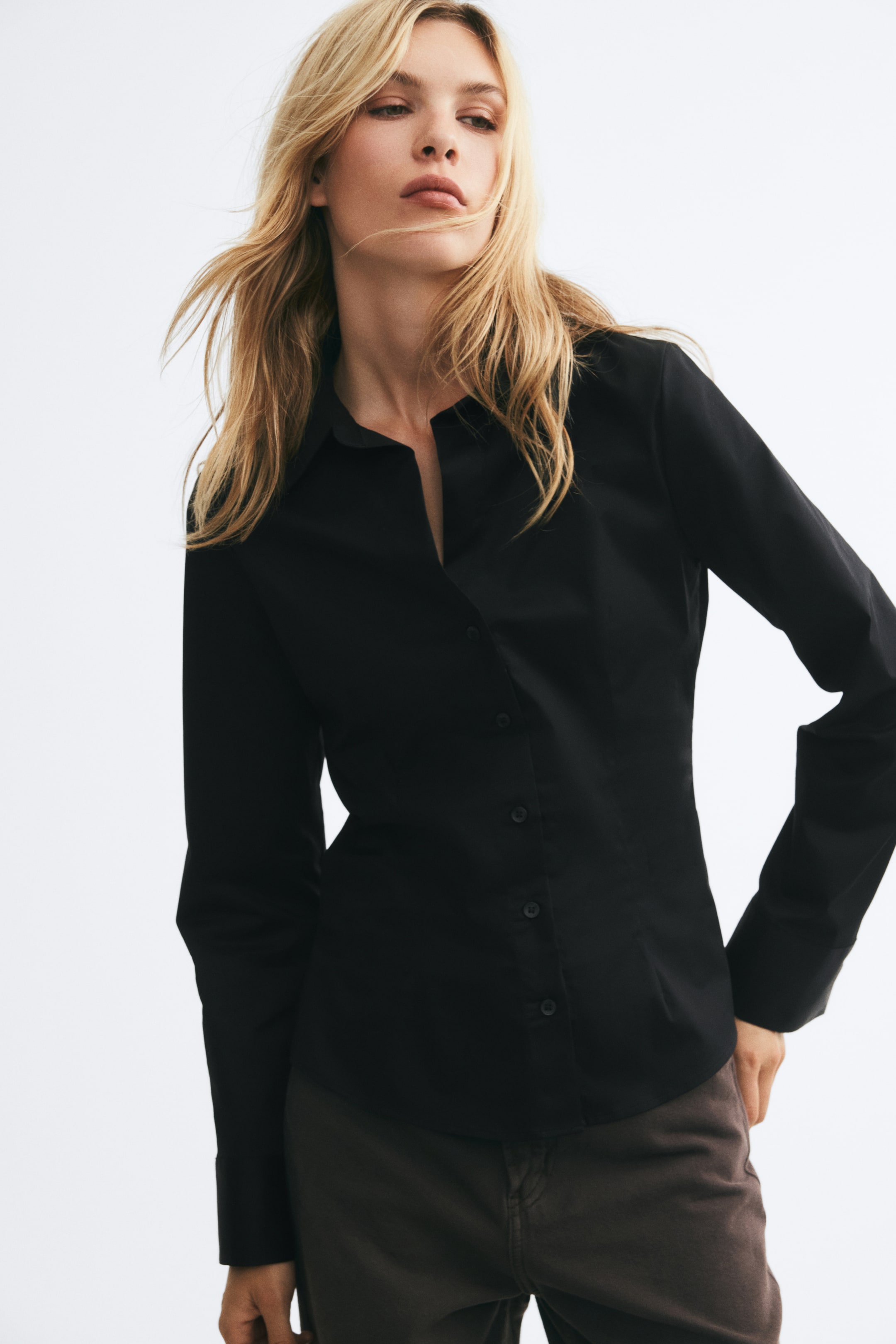 Ver imagen más grande: La modelo lleva una camisa negra ajustada de manga larga, con cuello abierto, cierre delantero de botones y pinzas de entalle sutiles. La camisa se combina con un pantalón marrón oscuro.