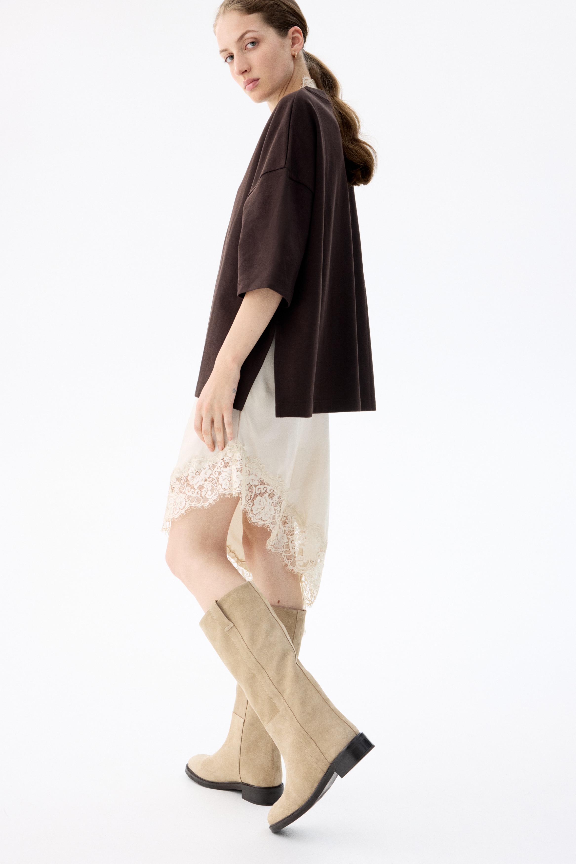 A single tan suede boot is standing in a side profile, featuring a rounded toe, a knee-high shaft with a stitched vertical strap detail, and a low black block heel.